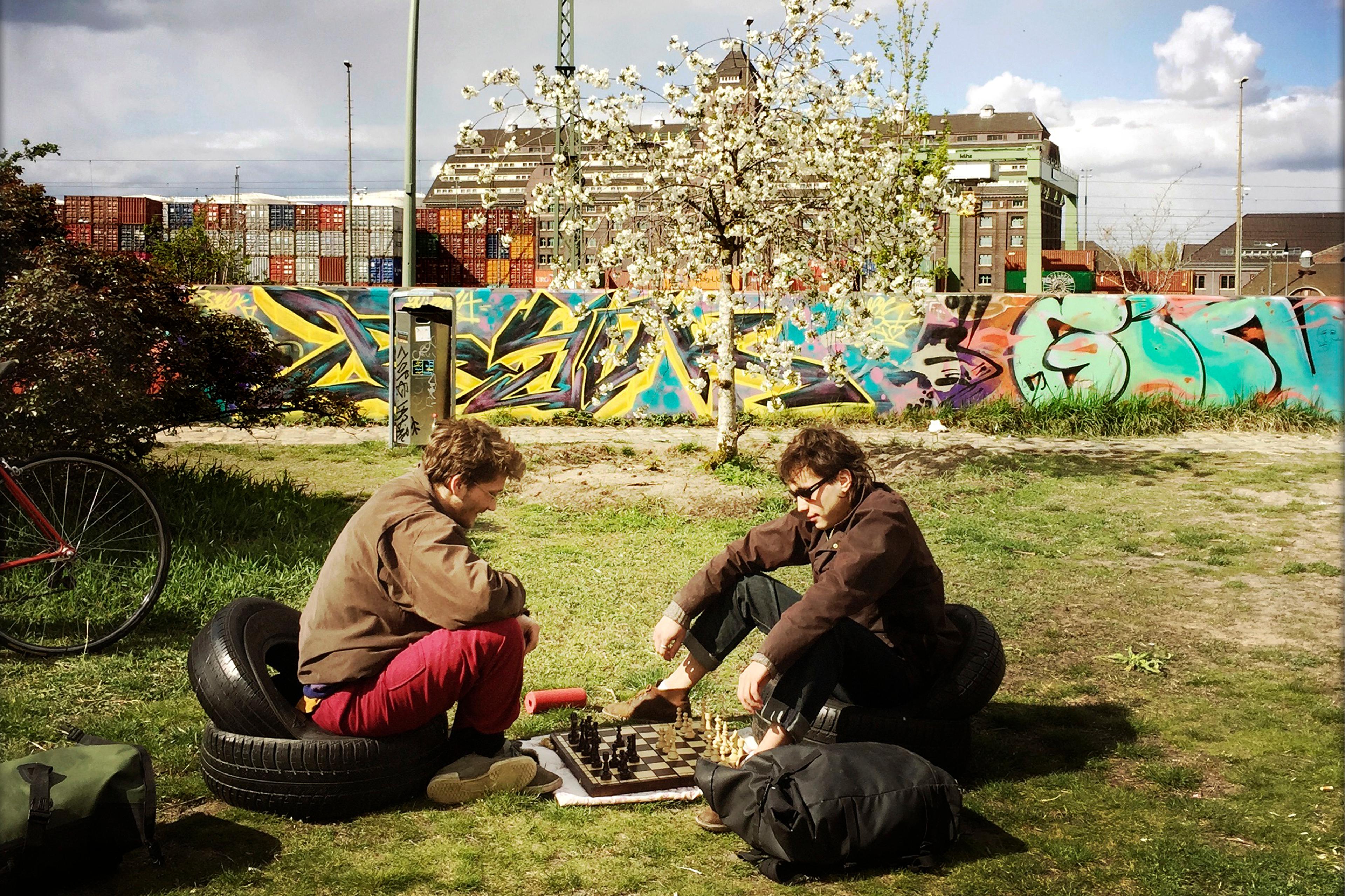 Photo of two people playing chess outdoors, seated on tyres near a graffiti-covered wall and blooming tree under cloudy skies.