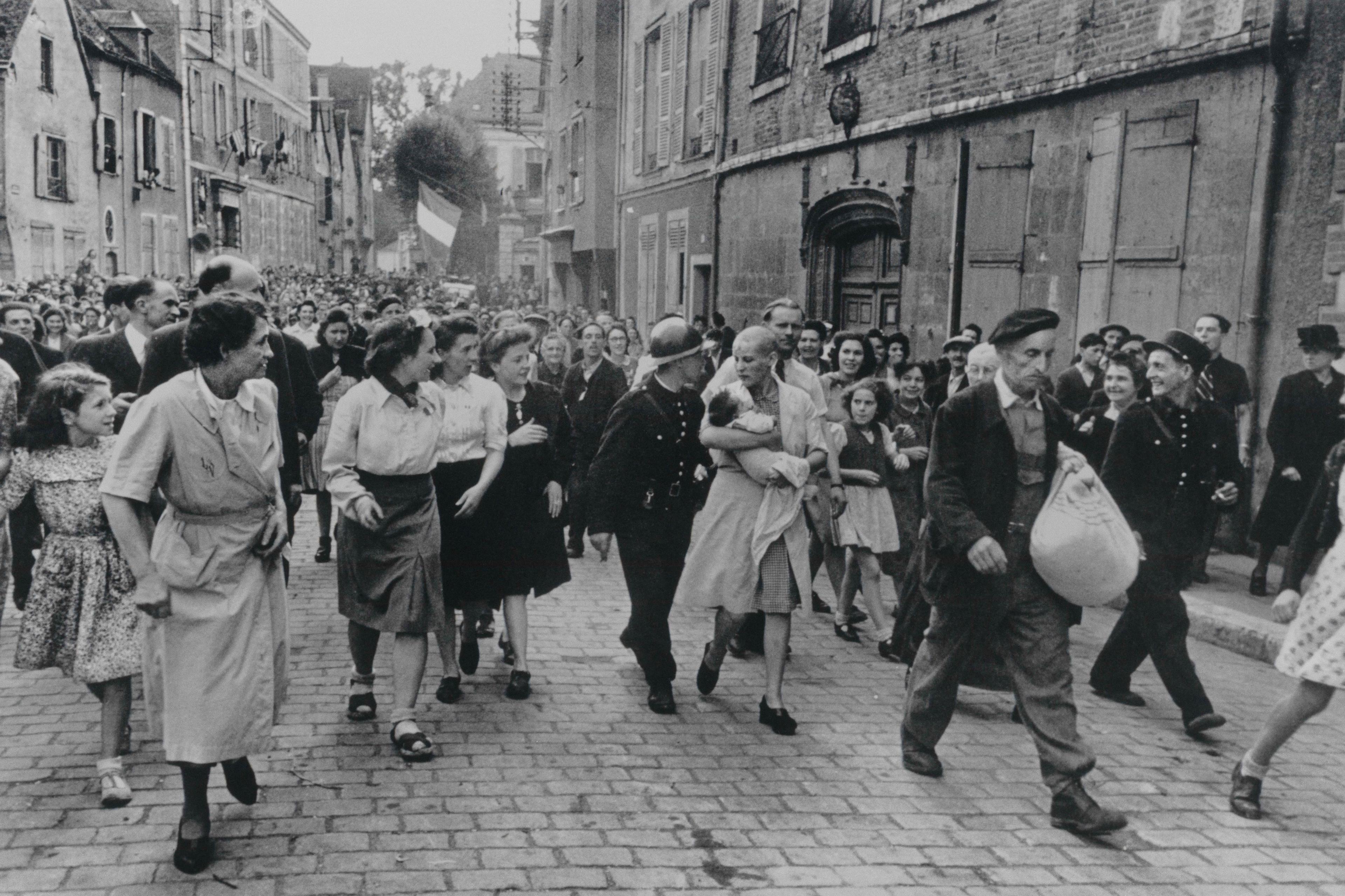 Black and white photo of a crowd of people walking down a cobblestone street with buildings in the background.