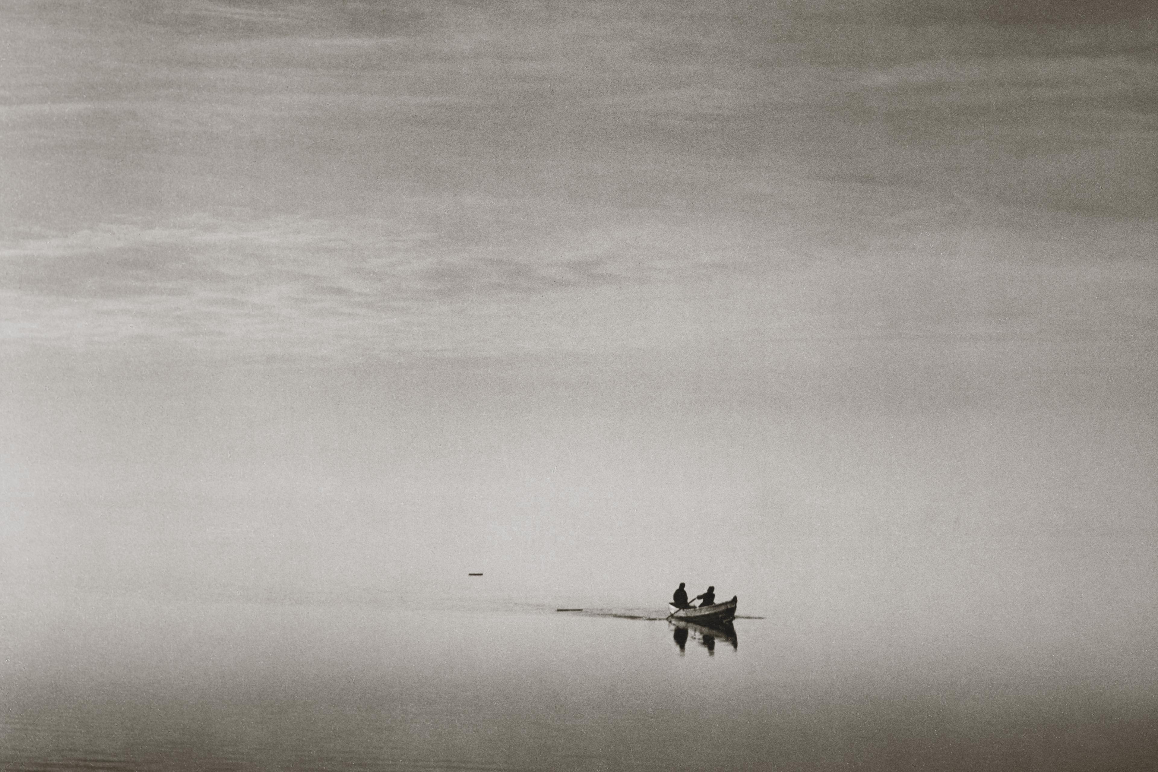 Black and white photo of a boat with two people on a calm lake, branches above and clouds reflected in the water.