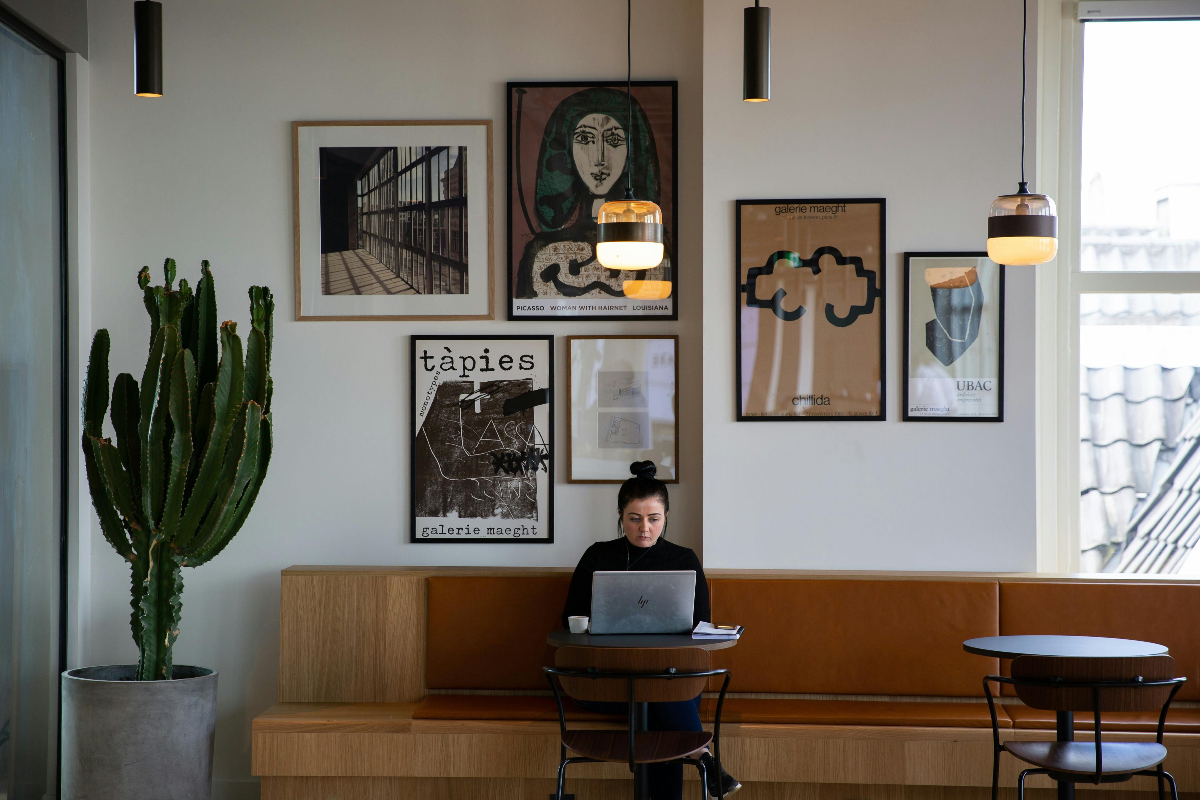 A woman on a laptop in a modern café with wall art, a large cactus and pendant lights above.