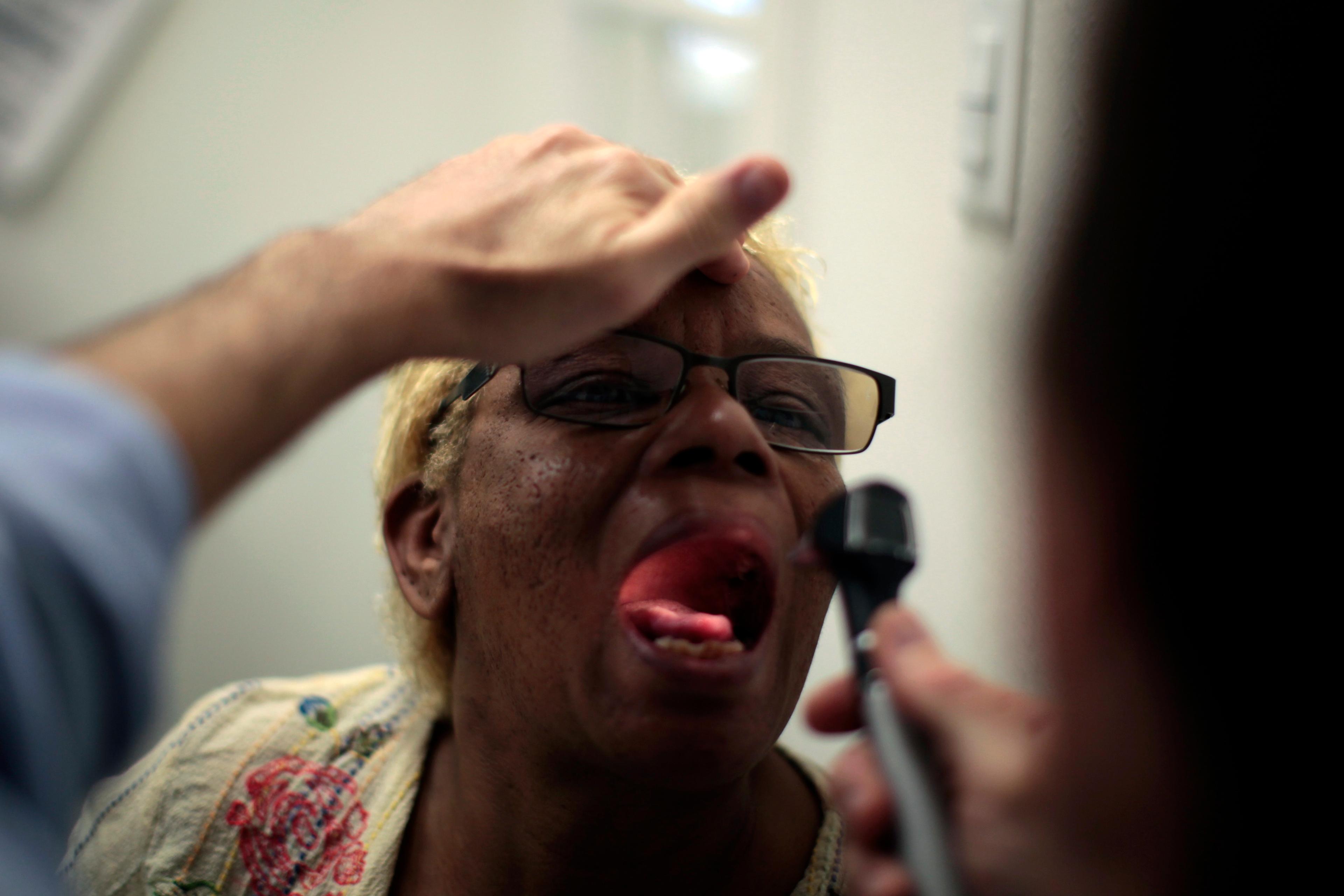 Photo of a person having their throat examined with an otoscope, wearing glasses and a patterned shirt, mouth open.