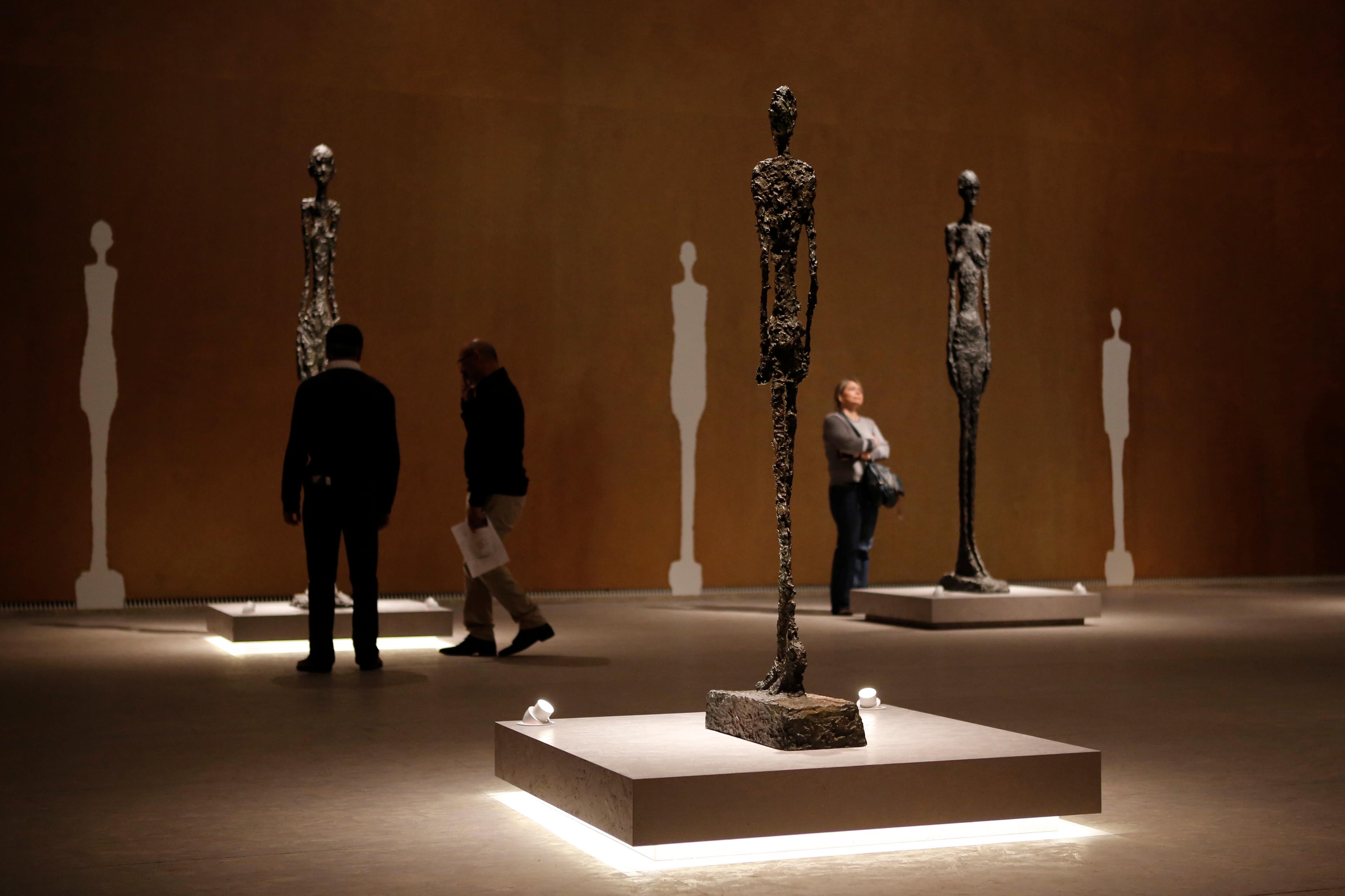 Photo of people viewing tall abstract sculptures in a dimly lit gallery with silhouetted artwork on the walls.