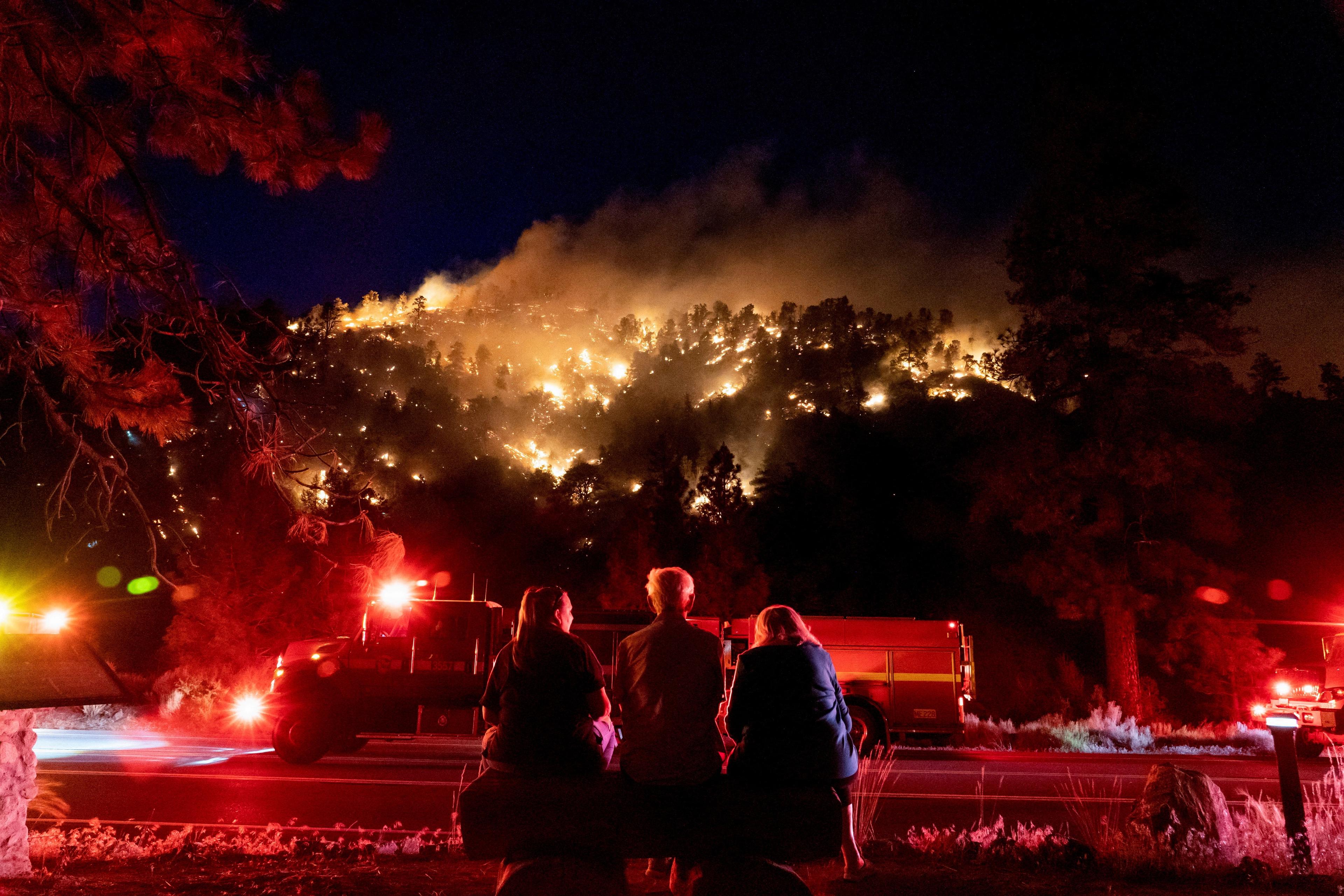 Photo of three people watching a wildfire on a hill at night with fire engines and flashing lights in the foreground.