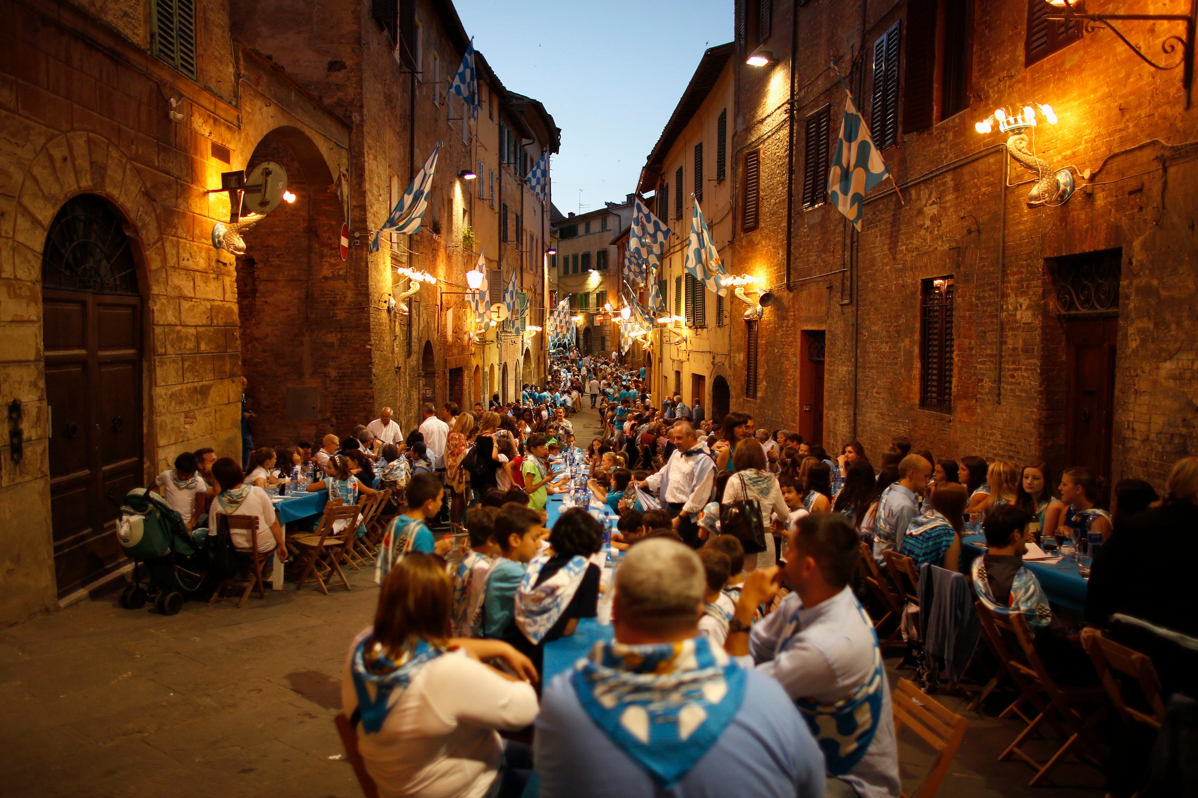 Photo of a crowded street dinner with blue flags, people sitting at long tables amid warm lights in a historic setting.