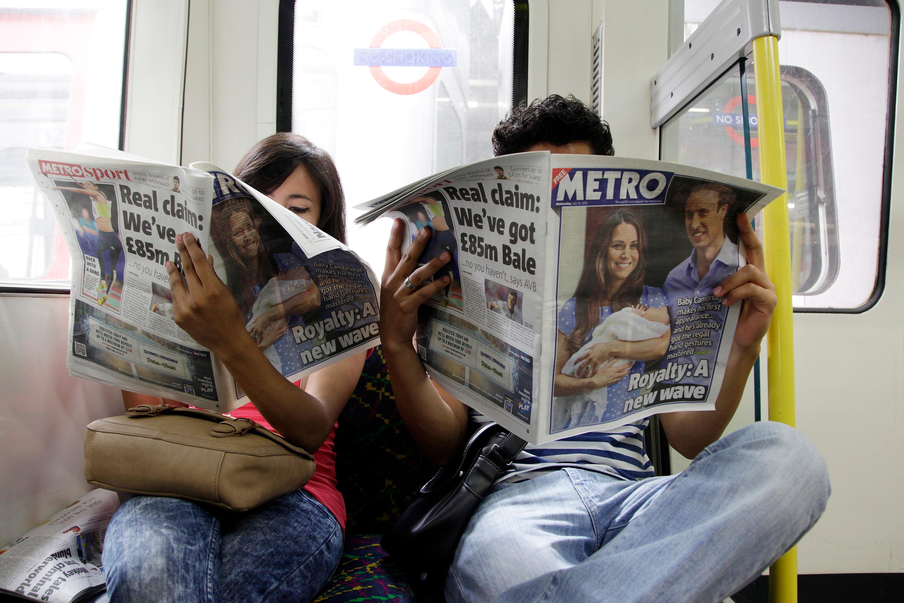 Two people on a train reading Metro newspapers with headlines about royalty and sports transfers.