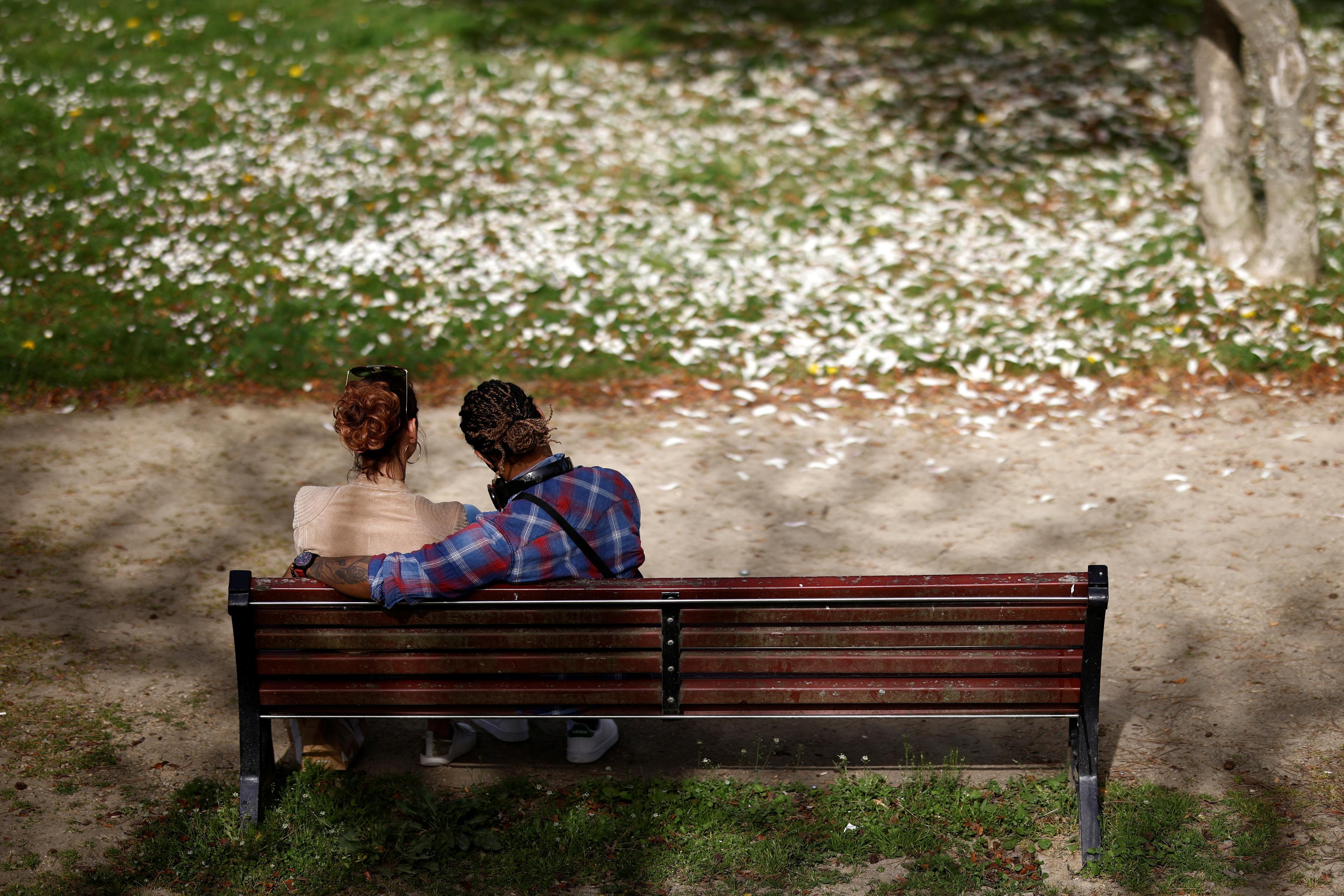Photo of two people sitting on a park bench, one with their arm around the other, surrounded by grass and fallen petals.