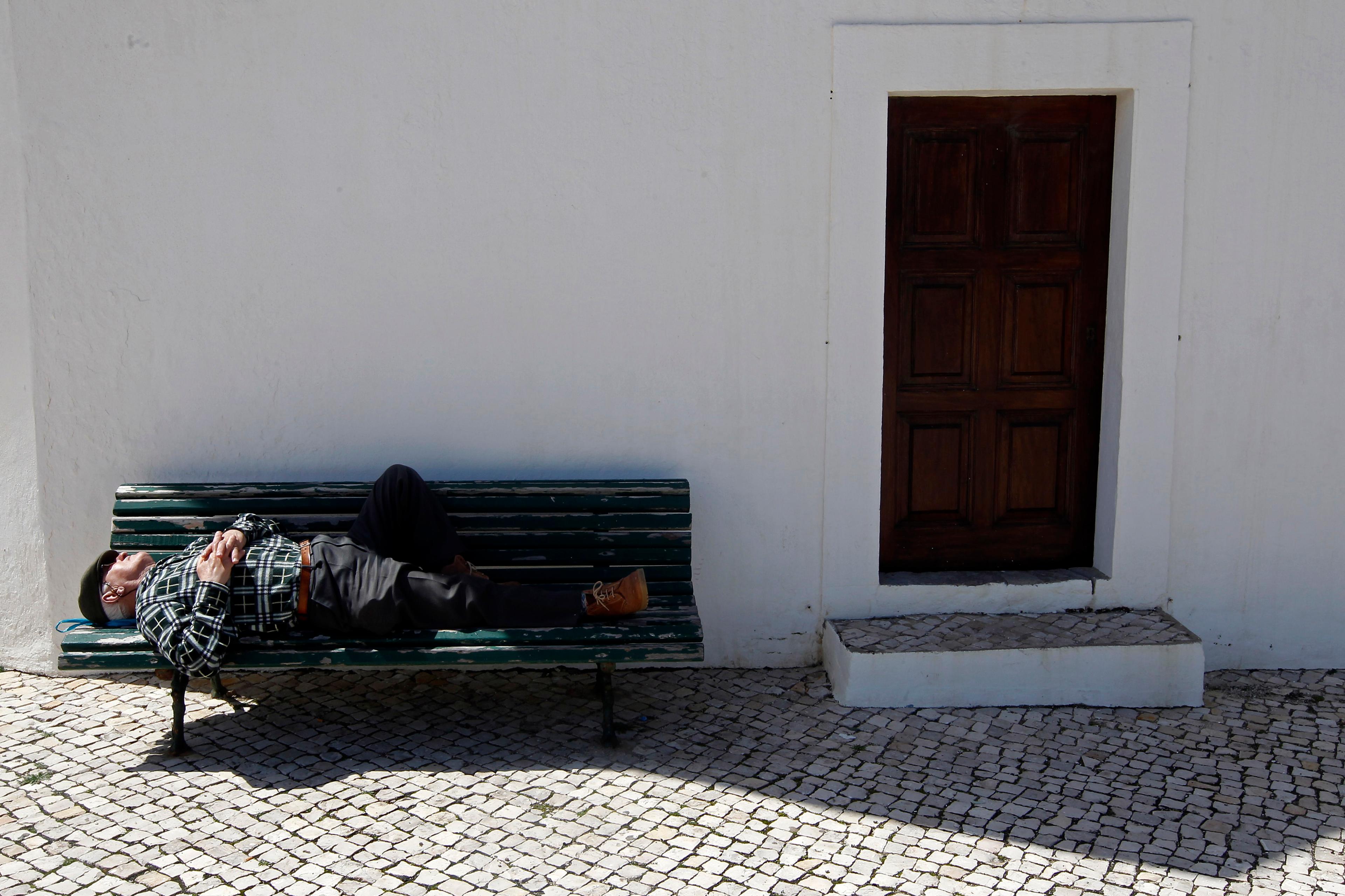 Photo of a man sleeping on a bench against a white wall near a wooden door, with cobblestone paving below in sunlight.