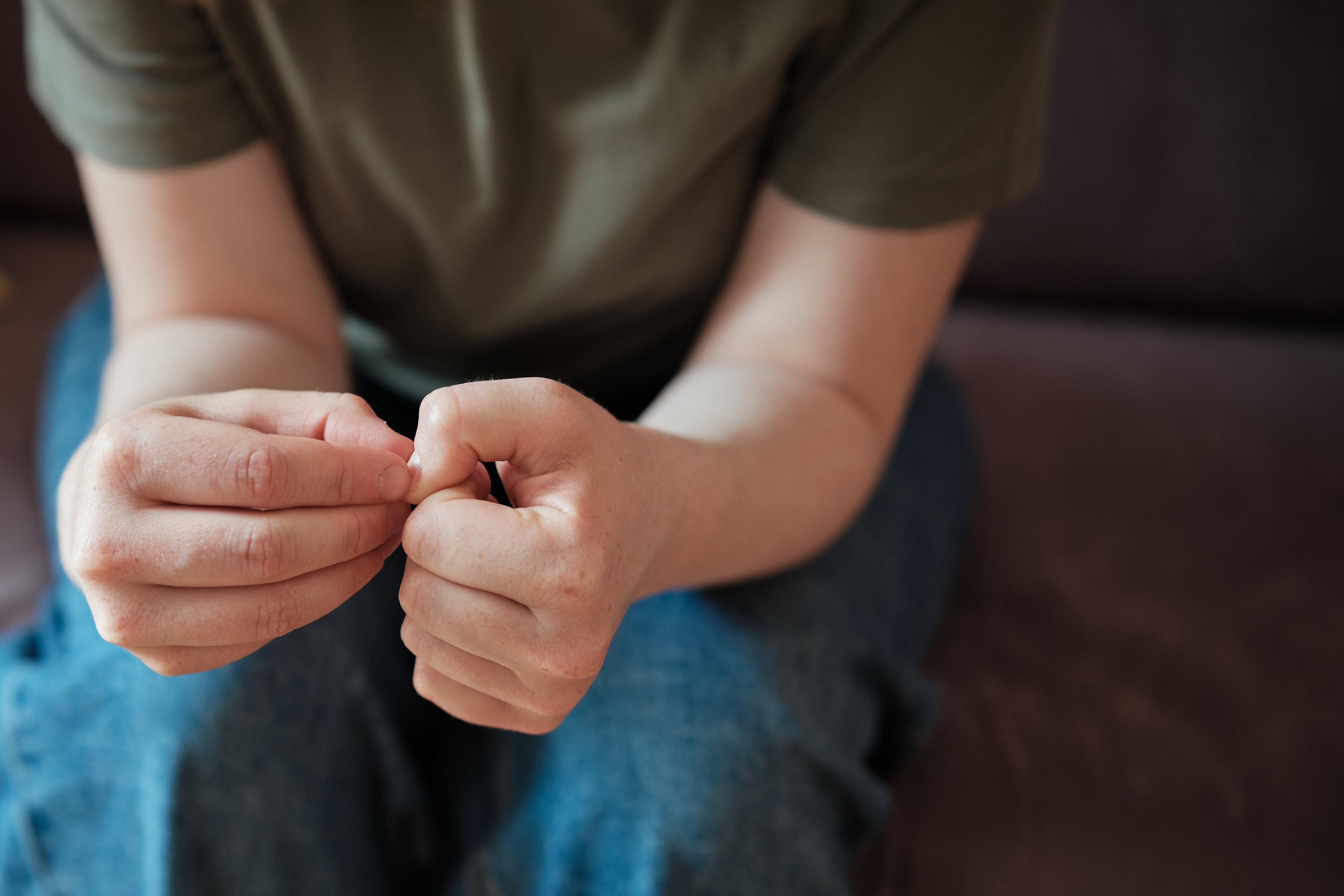A person in a green shirt and jeans sitting, focused on their hands which are clasped together, the right hand picking at the left hand’s thumbnail.