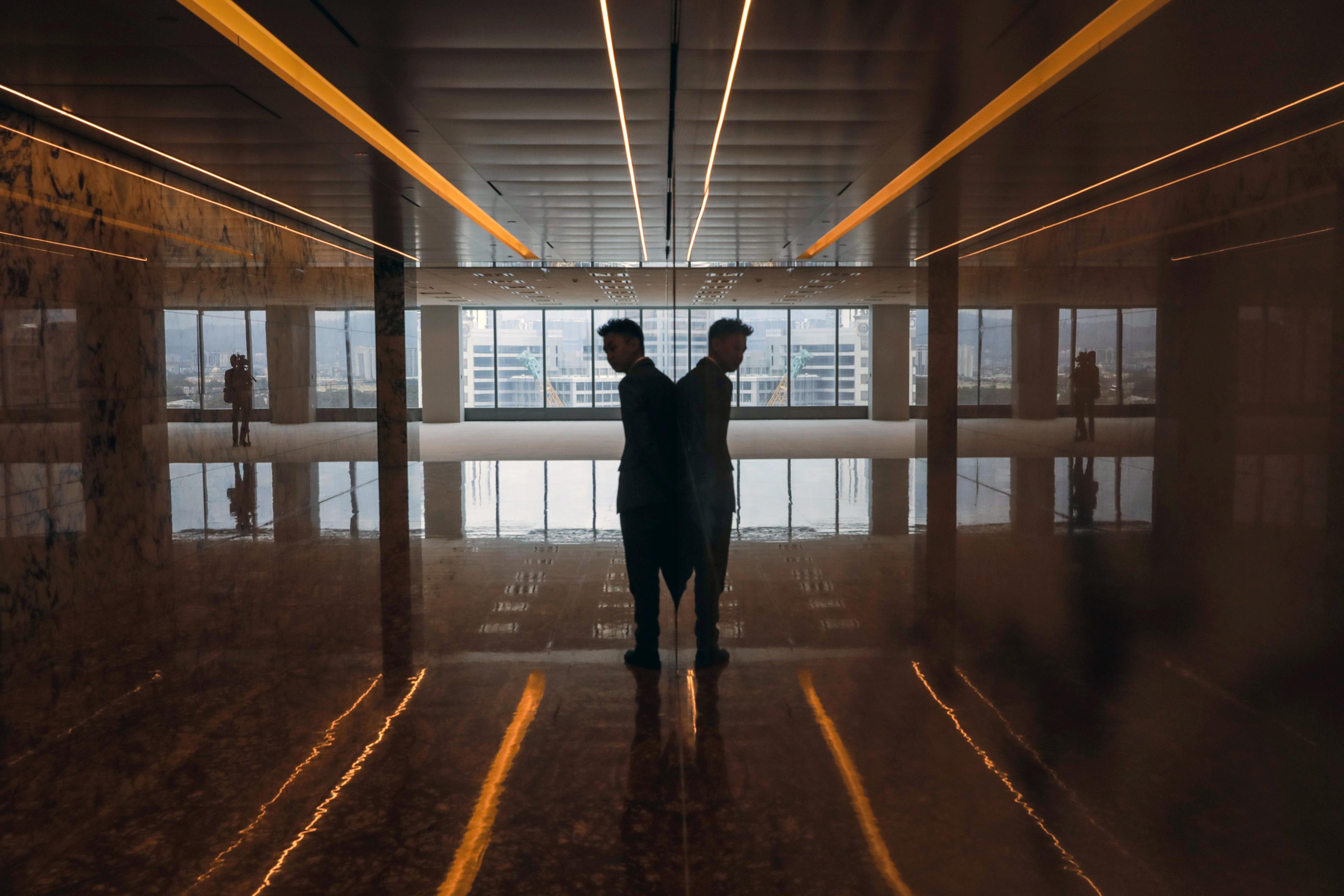 A man in silhouette standing against a reflective surface in an empty room with cityscape visible through large windows.
