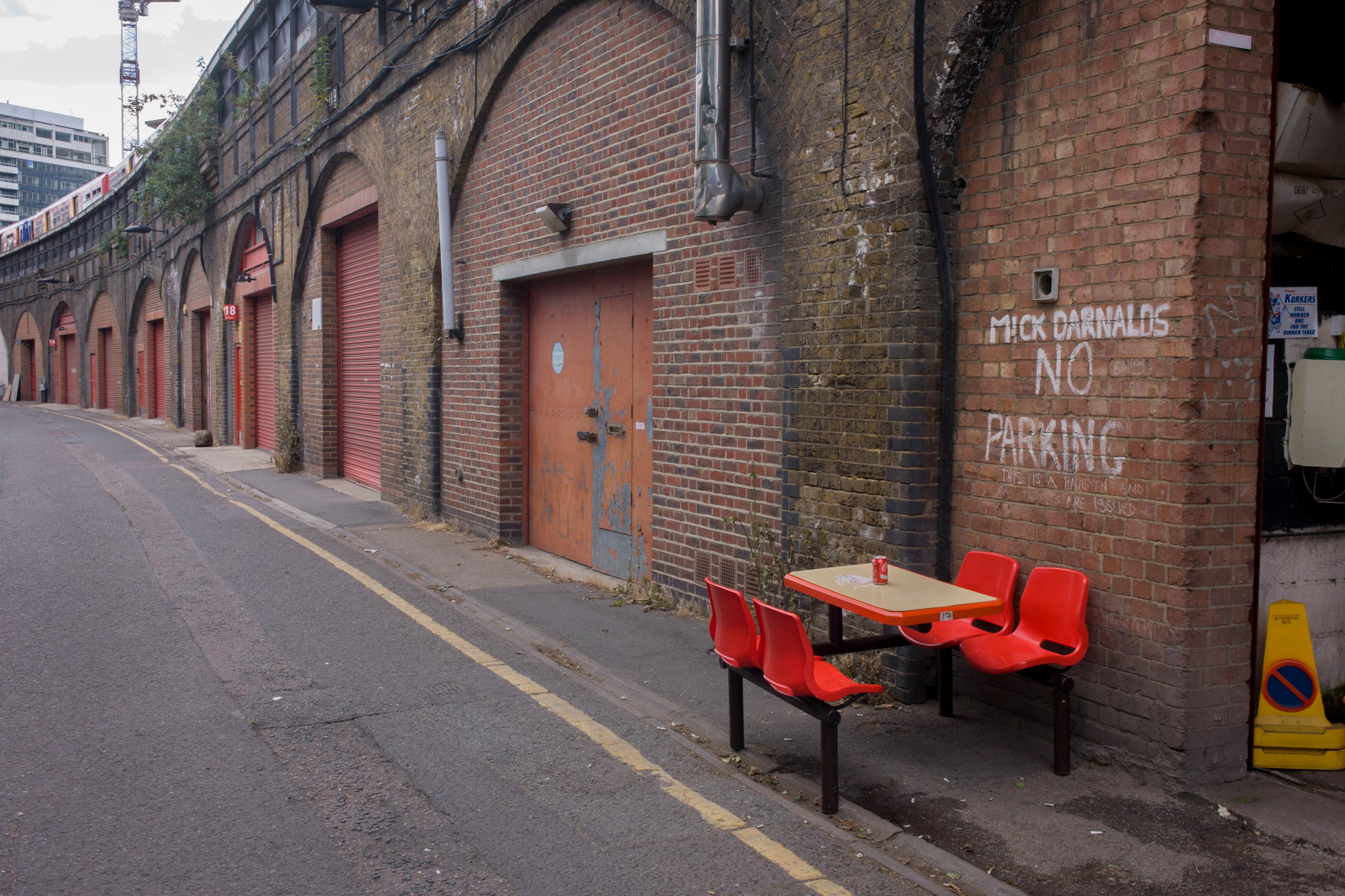A road below a railway line with a brick wall and red garage doors, graffiti that reads ‘MICK DARNALDS NO PARKING’ behind a table with a can of coke on it and four red chairs.