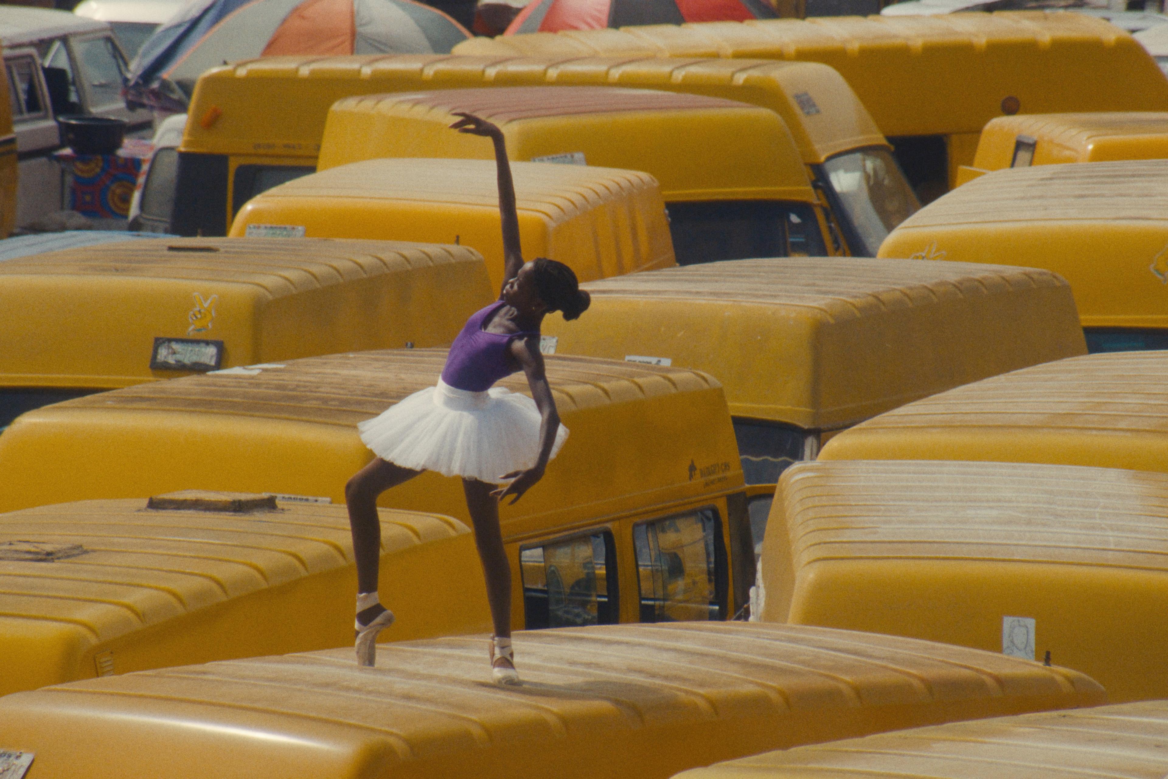 A Nigerian ballerina in a white tutu dancing atop yellow minibuses in a crowded urban setting.