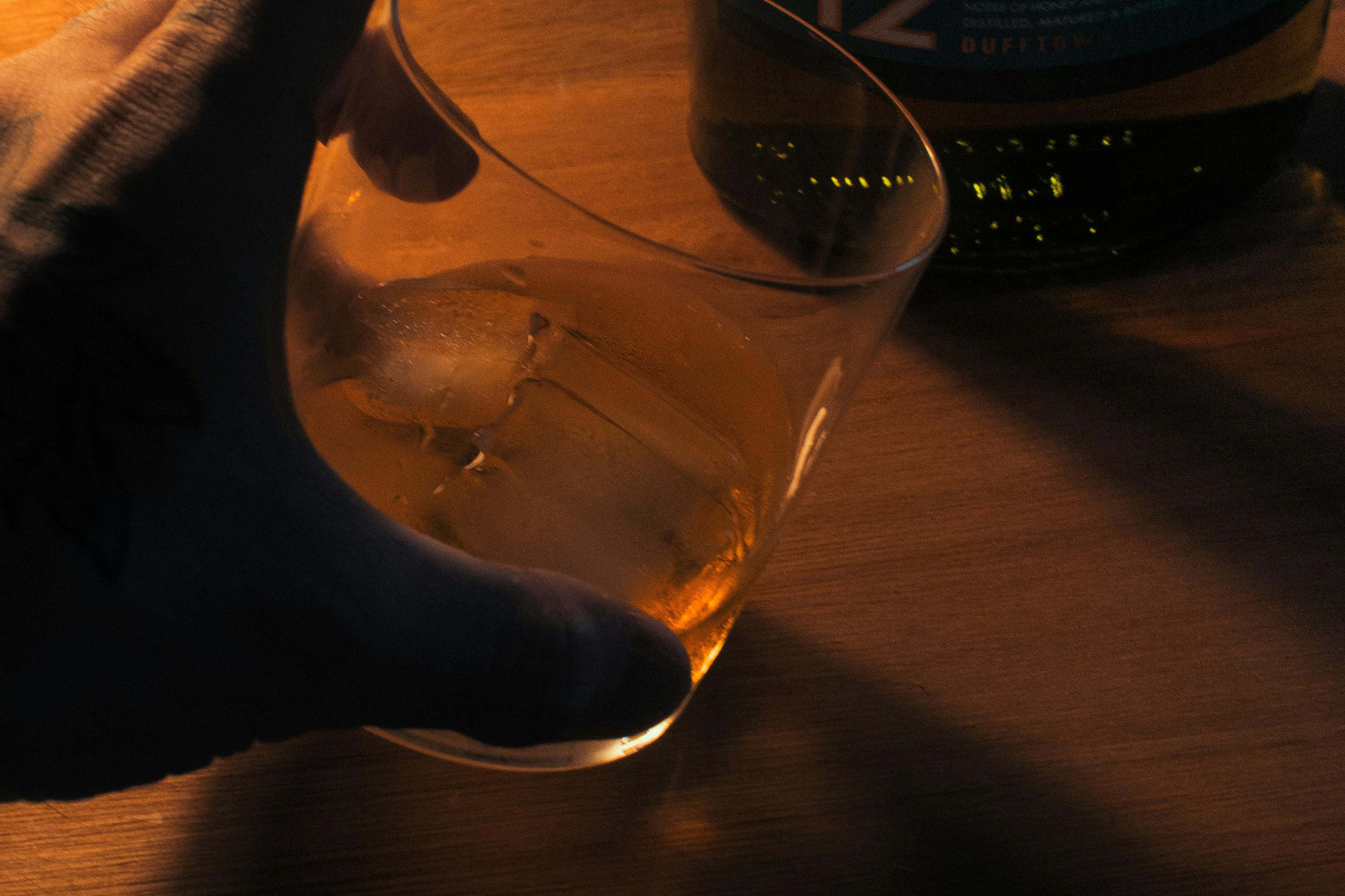 Photo of a hand holding a glass of whisky with ice next to a bottle on a wooden surface, dimly lit setting.