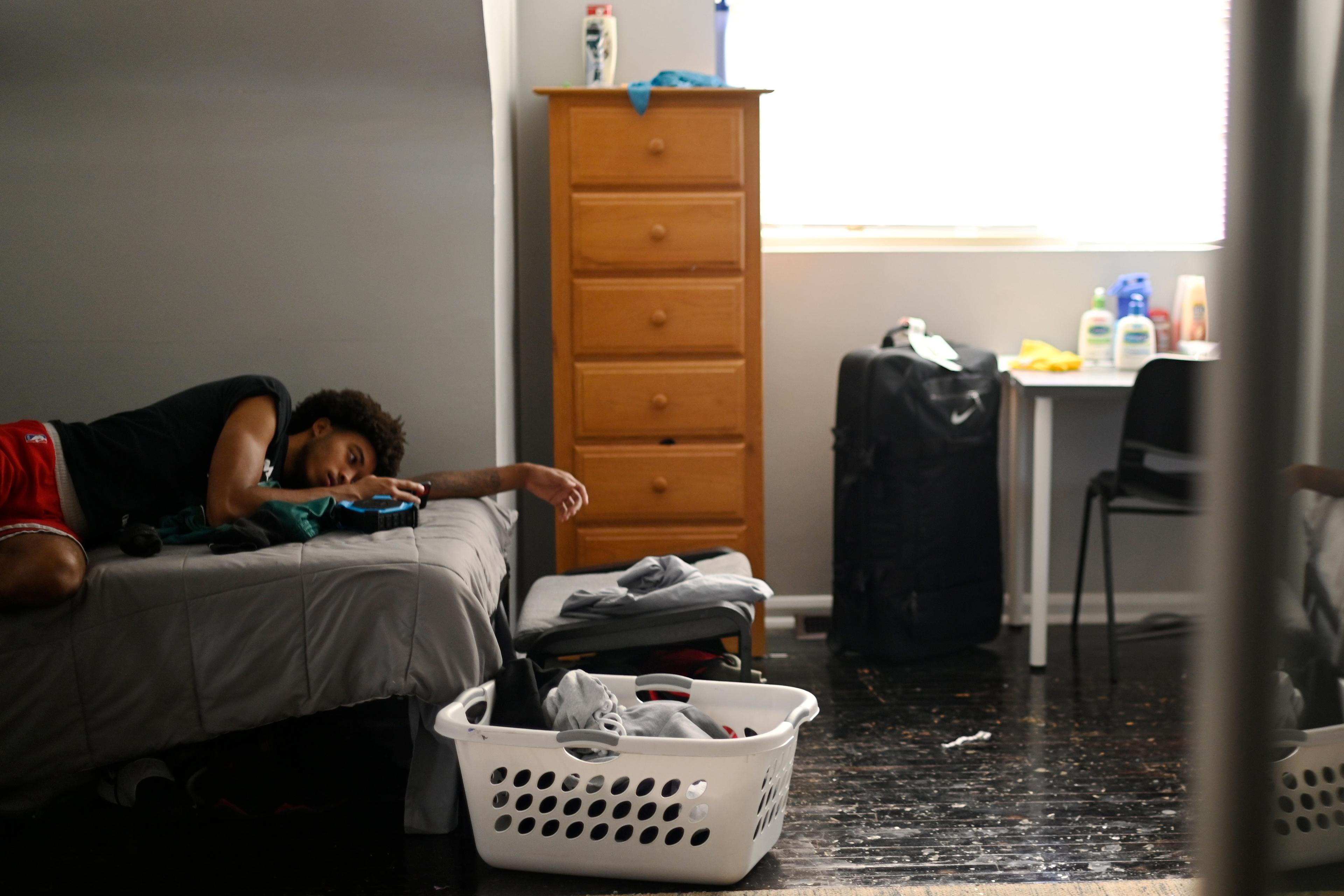 A young person lying on a bed in a cluttered room with a suitcase, laundry basket, chest of drawers and desk, illuminated by window.