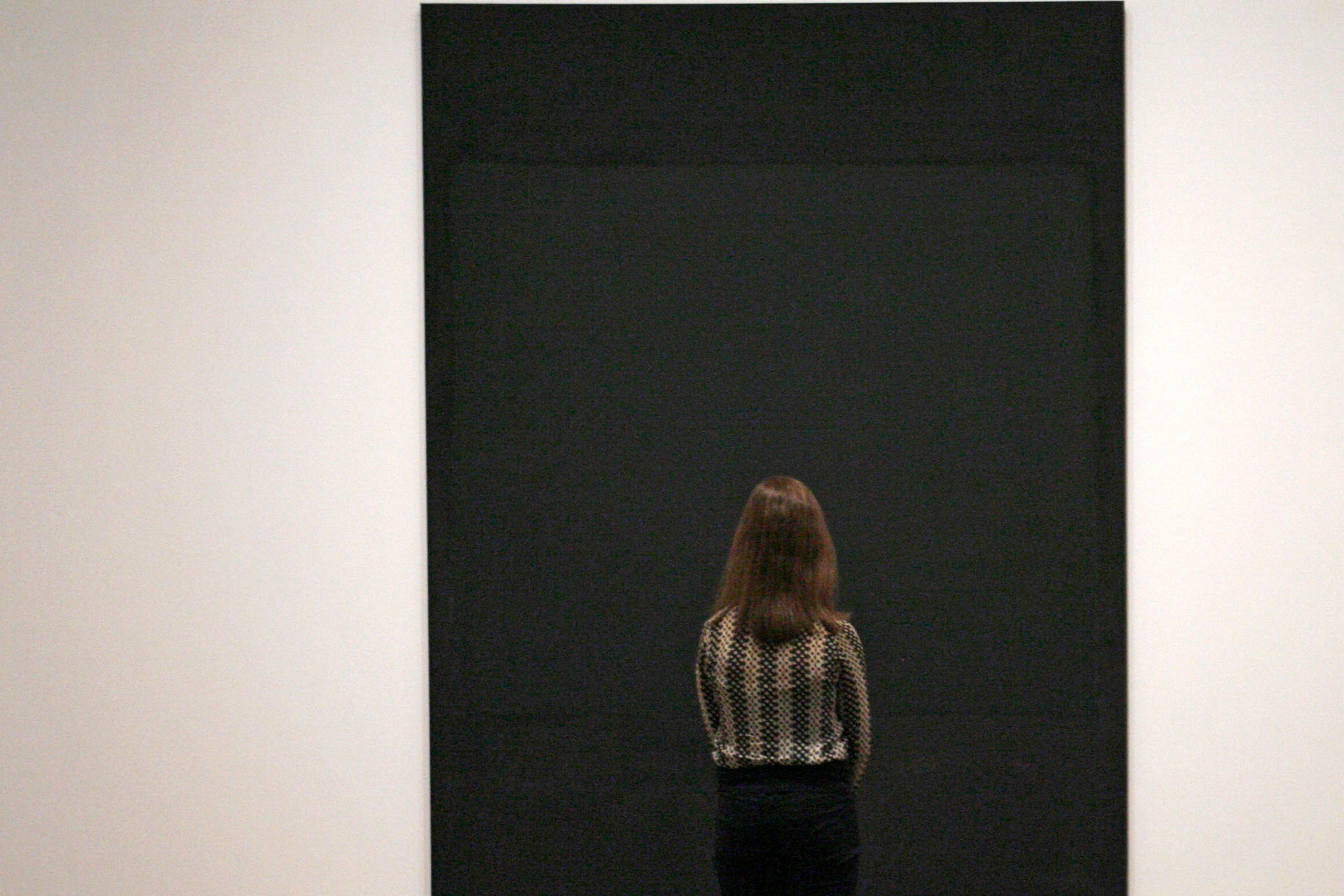 Photo of a woman viewed from behind looking at a large black abstract painting in a minimalist gallery setting.