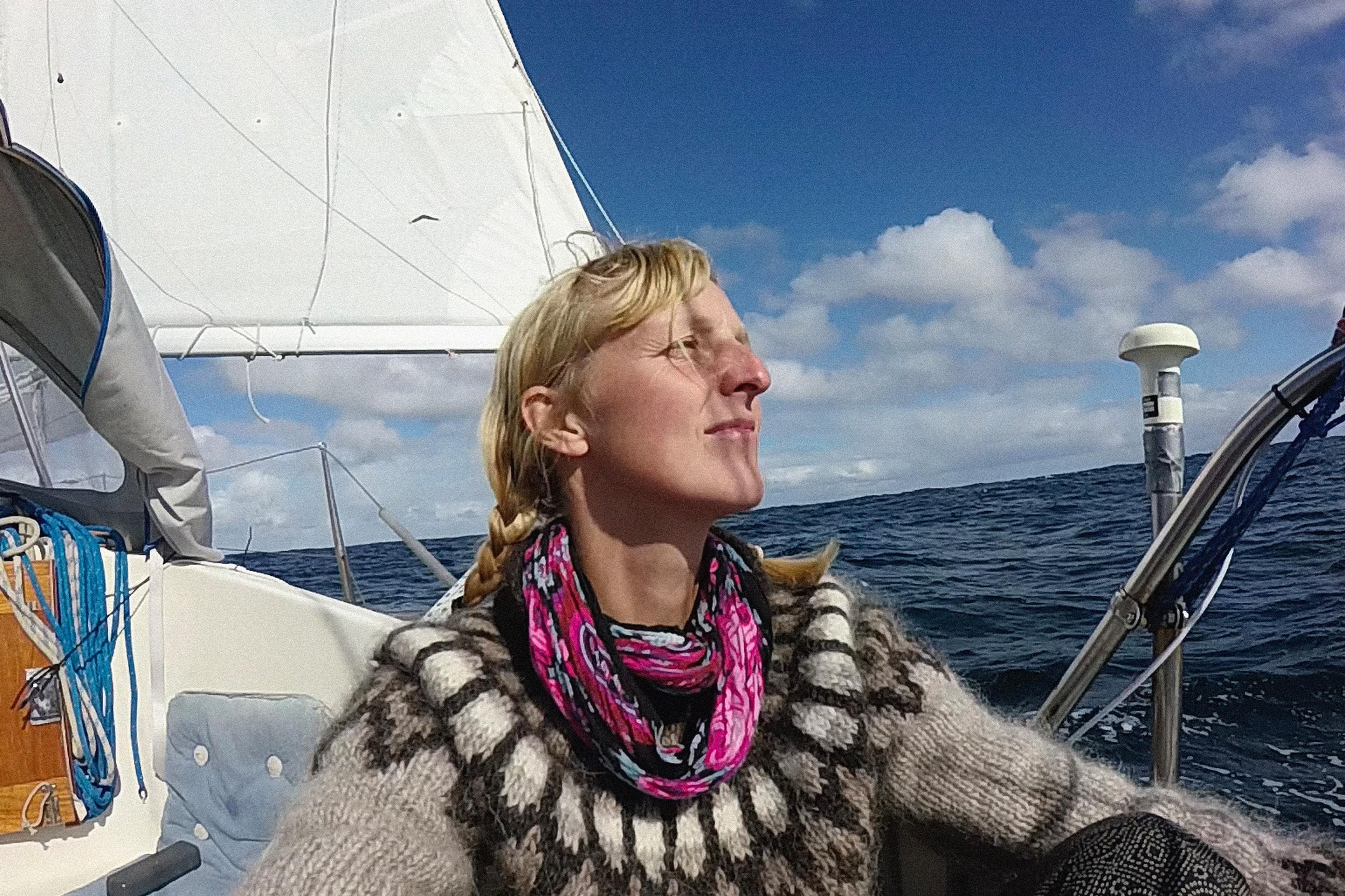 Photo of a woman with braided hair on a sailboat in open sea, wearing a patterned jumper and colourful scarf, enjoying the sun.