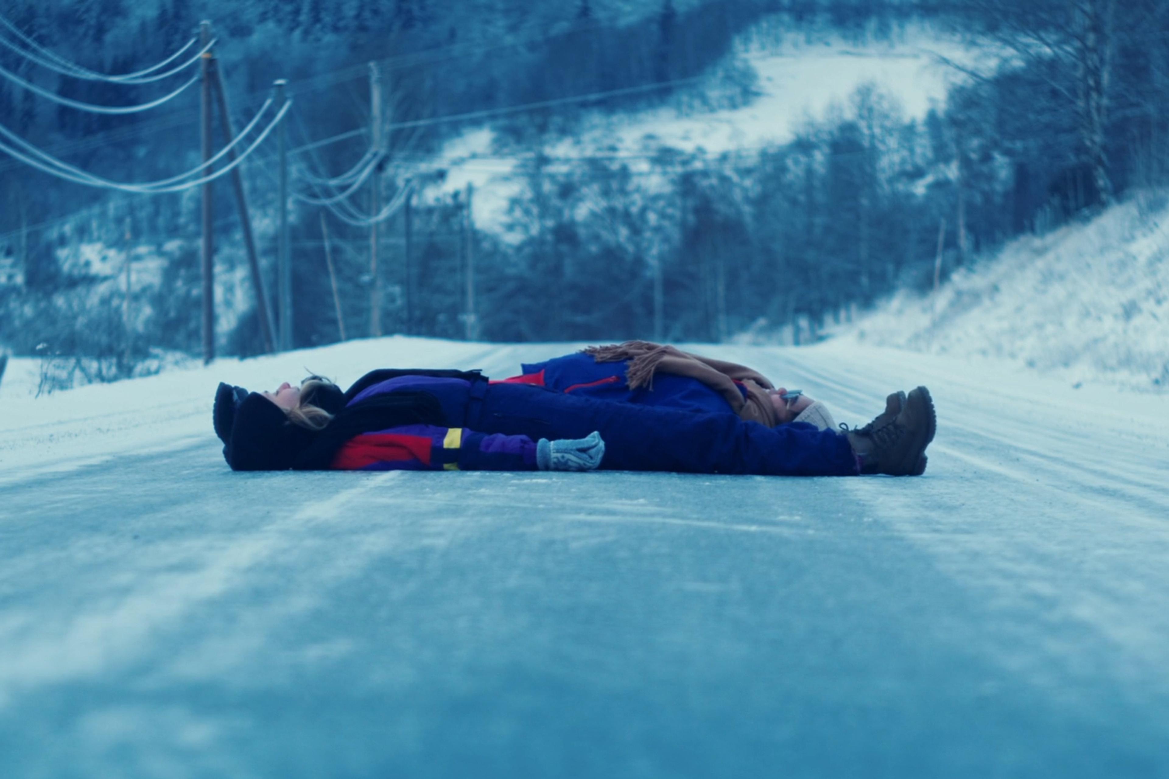 Two people lying on a snowy road in winter clothing with a forested landscape in the background.
