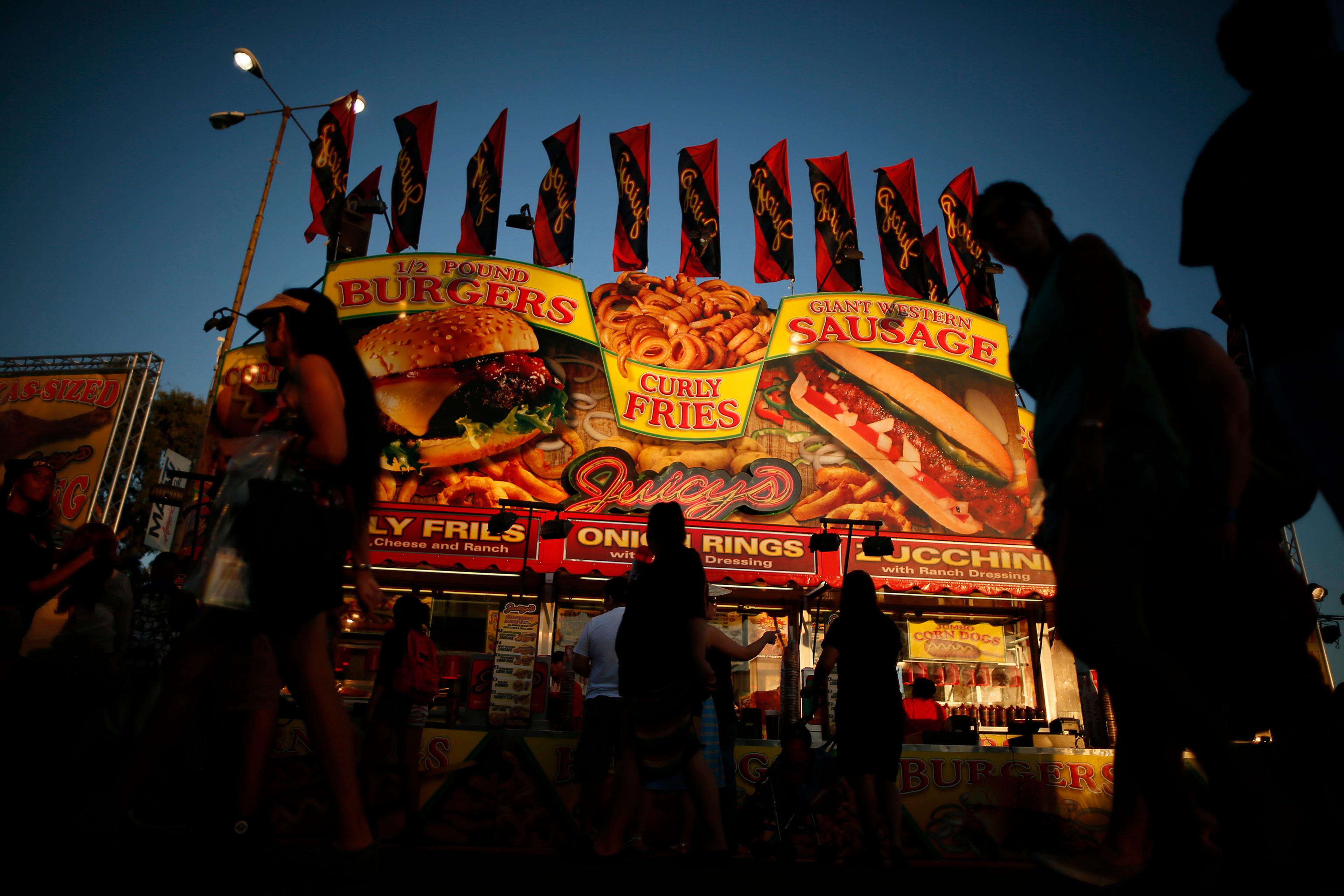 A bustling food stall at dusk with bright signs advertising burgers, curly fries and sausages against a darkening sky.