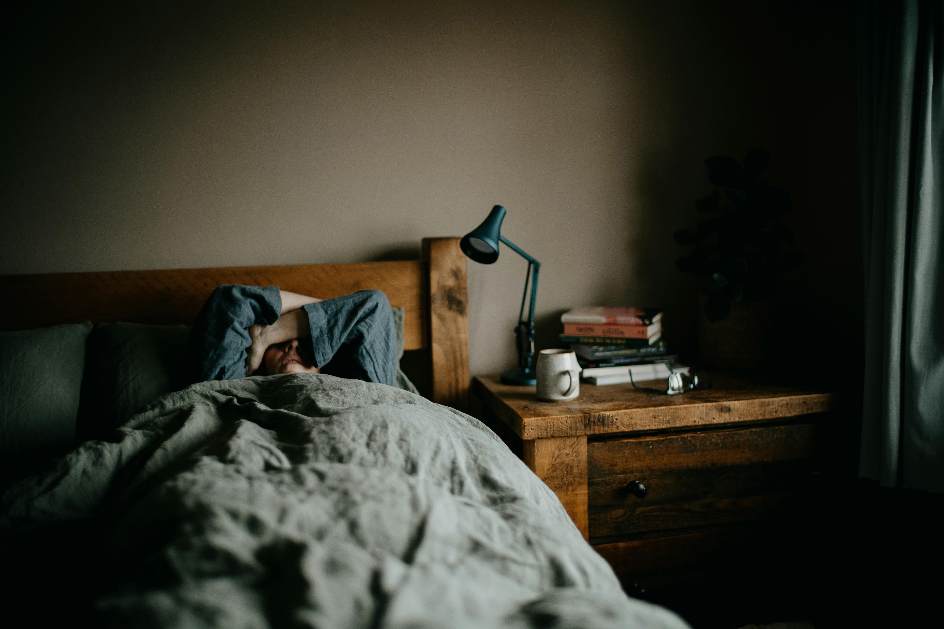 A person lying in bed under a duvet, arms covering face; bedside table with books, a lamp, a mug and reading glasses.