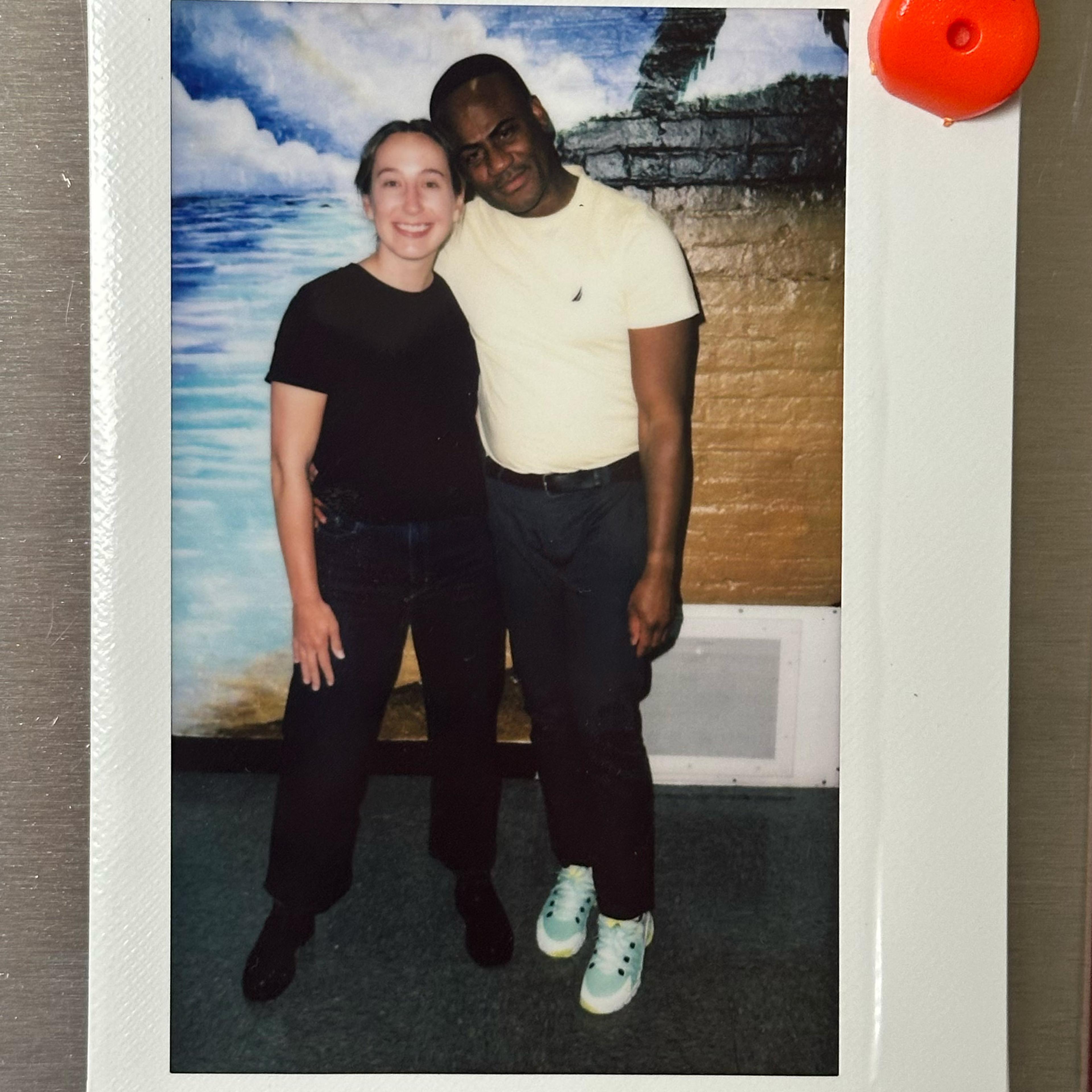 A Polaroid photo of two people standing together smiling in front of a painted mural depicting the sea and clouds.