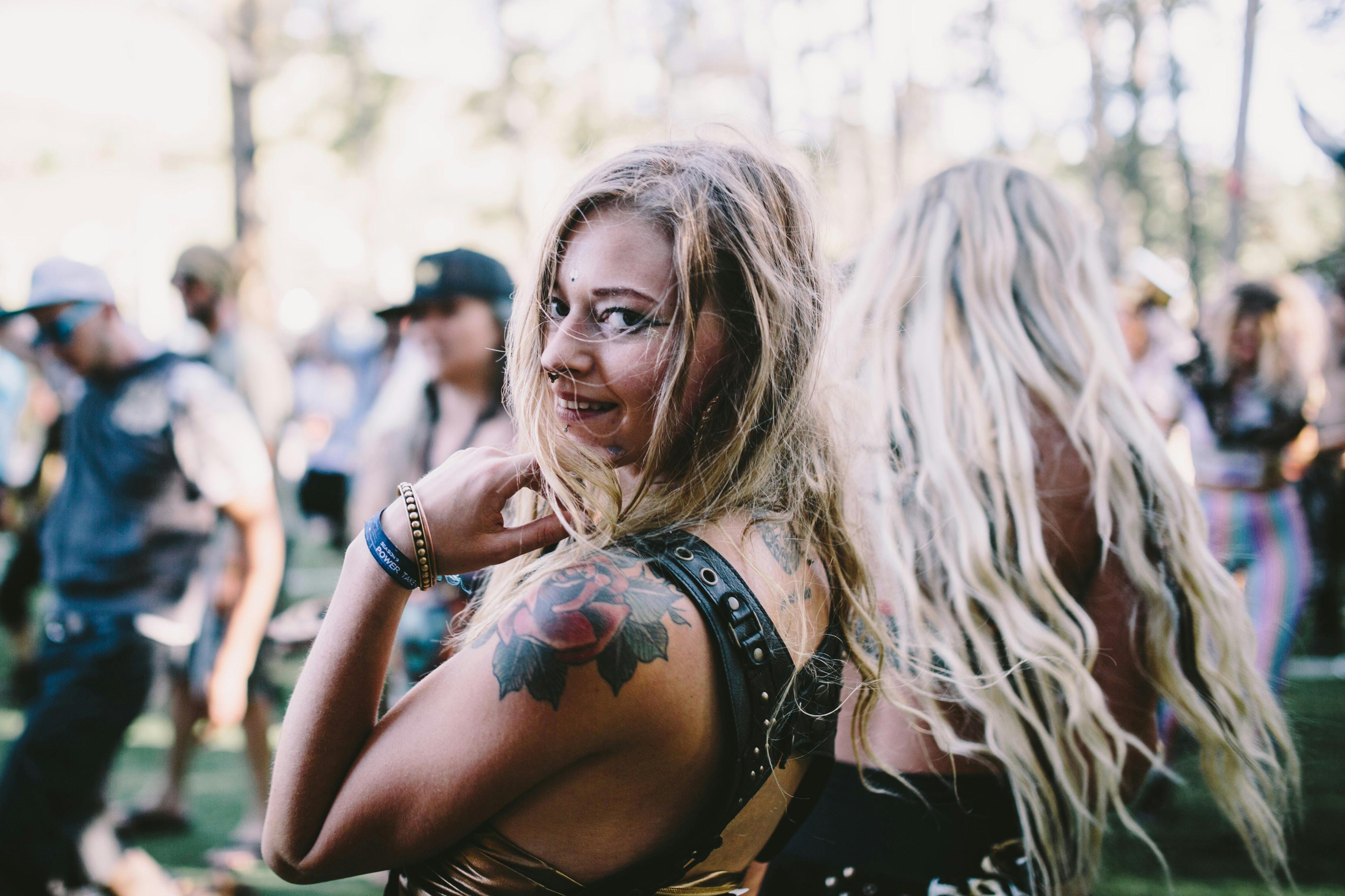 A smiling woman with tattoos at an outdoor festival, surrounded by blurred people and trees in the background.