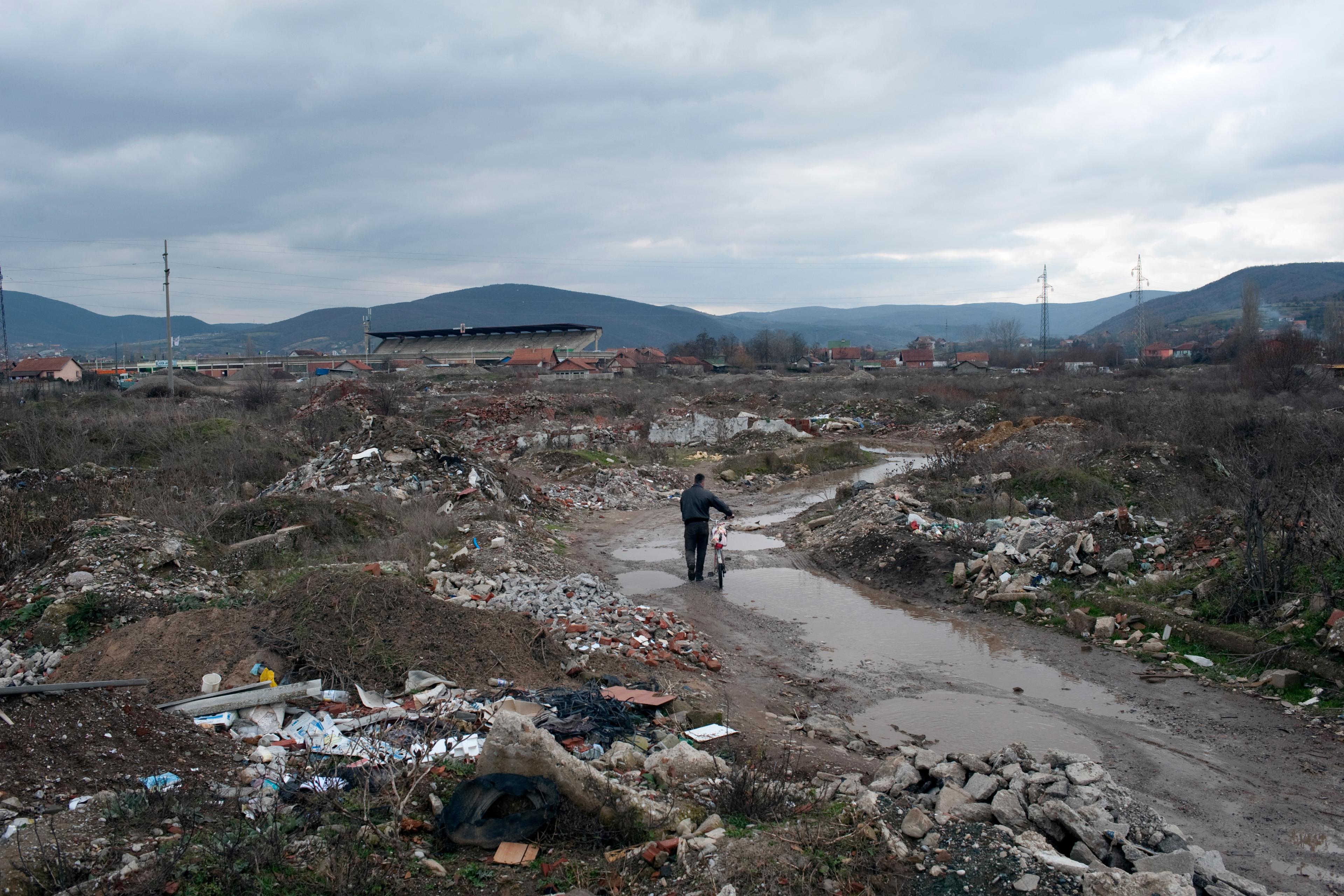 A person pushing a bike through a muddy landfill with debris under an overcast sky, distant hills and houses.