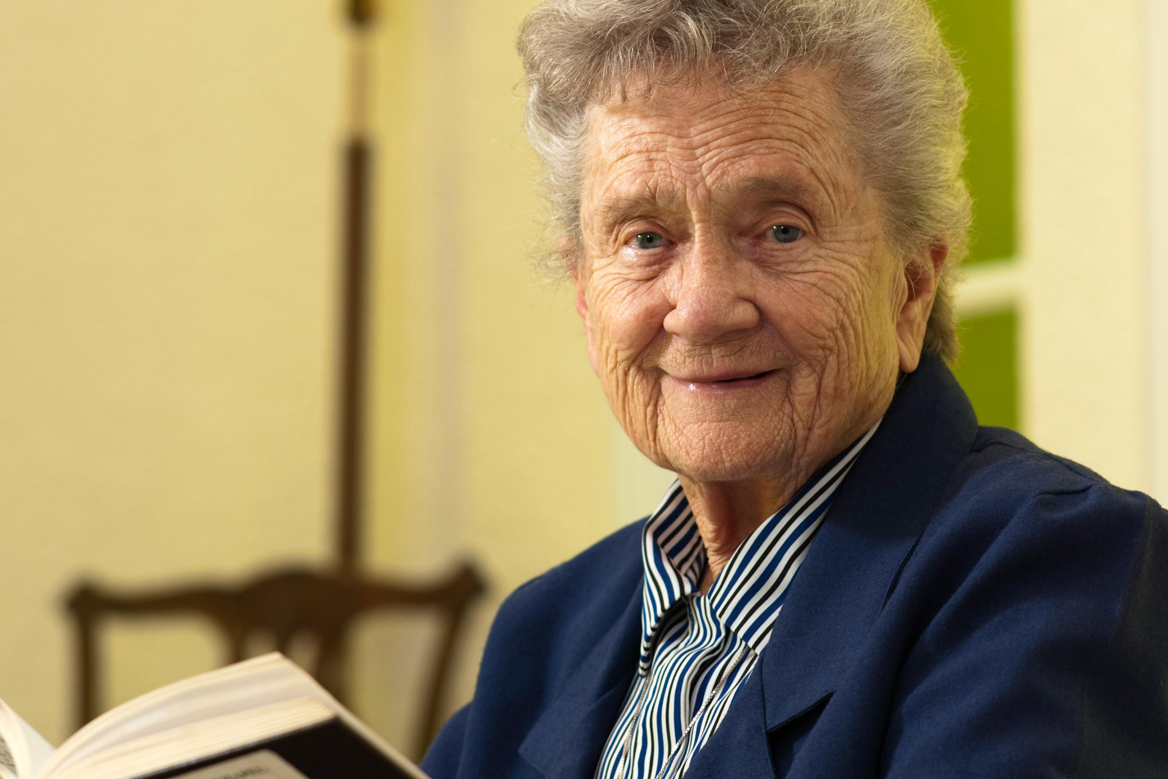 An elderly woman in a blue jacket holding a book, smiling warmly at the camera indoors.
