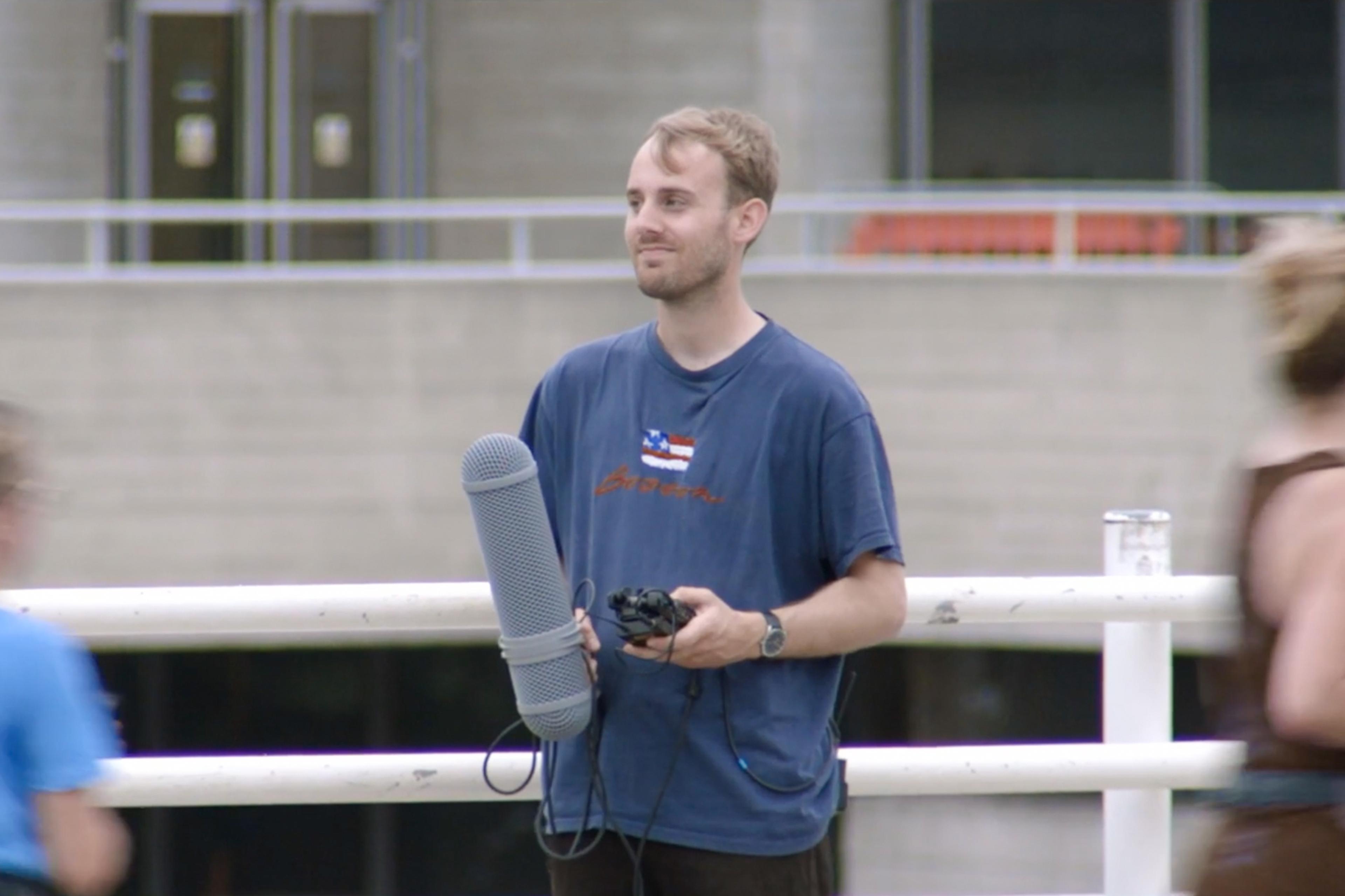 Photo of a man holding a microphone and equipment standing on a walkway with blurred people passing by in the foreground.