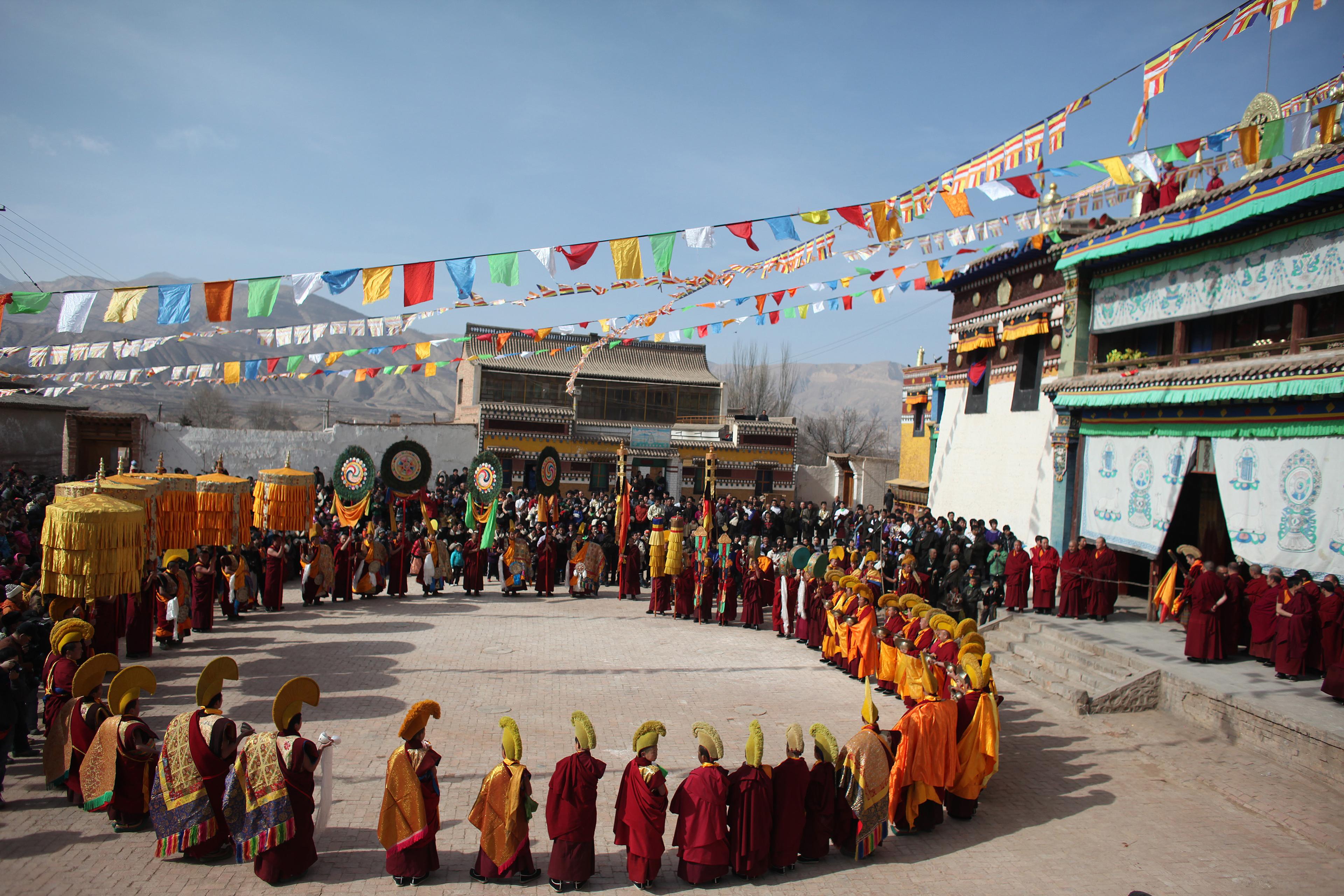 Monks in colourful robes and hats, forming a circle during an outdoor festival with flags and onlookers in Tibet.
