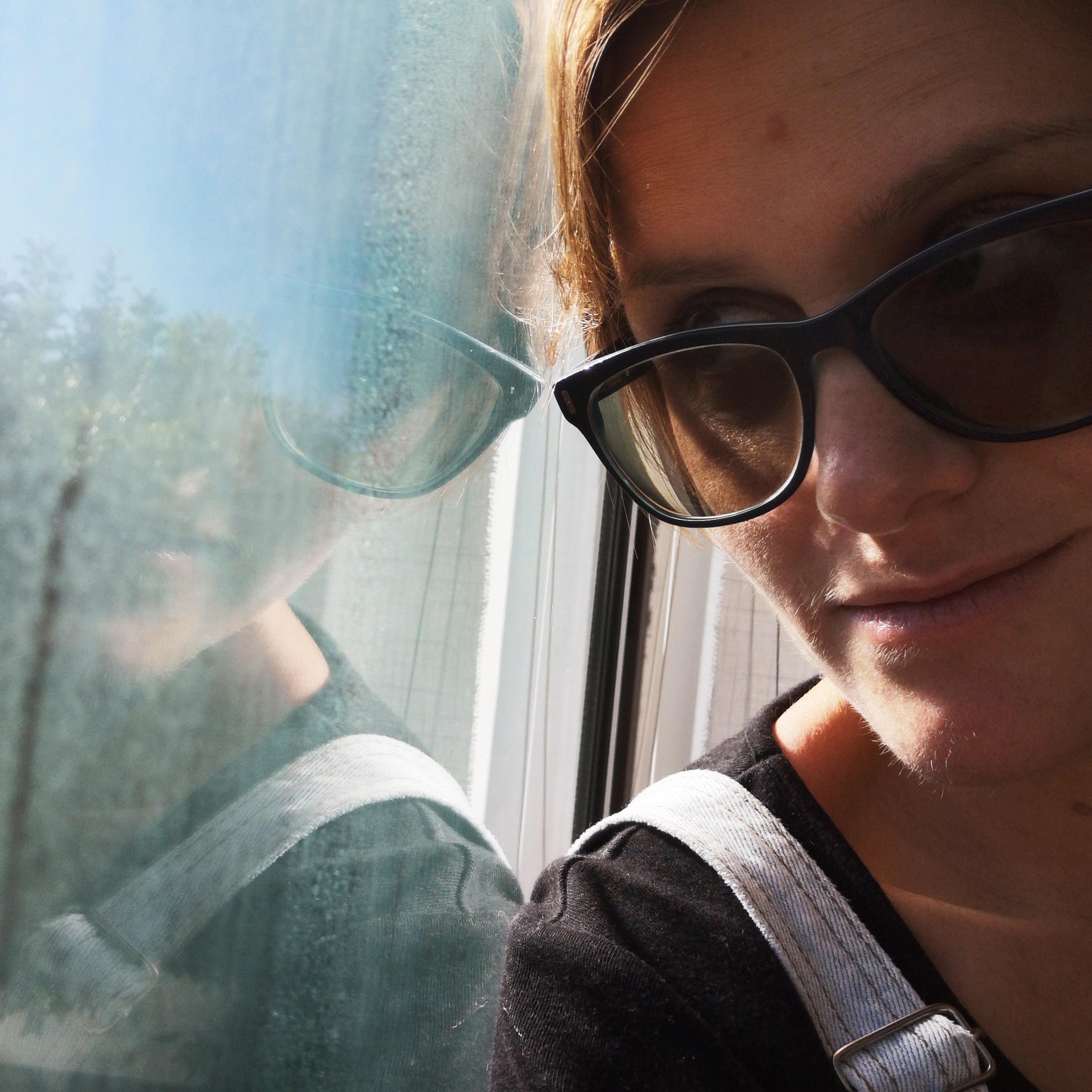 A person wearing sunglasses looking at their reflection in a window with a blurry outdoor scene visible.