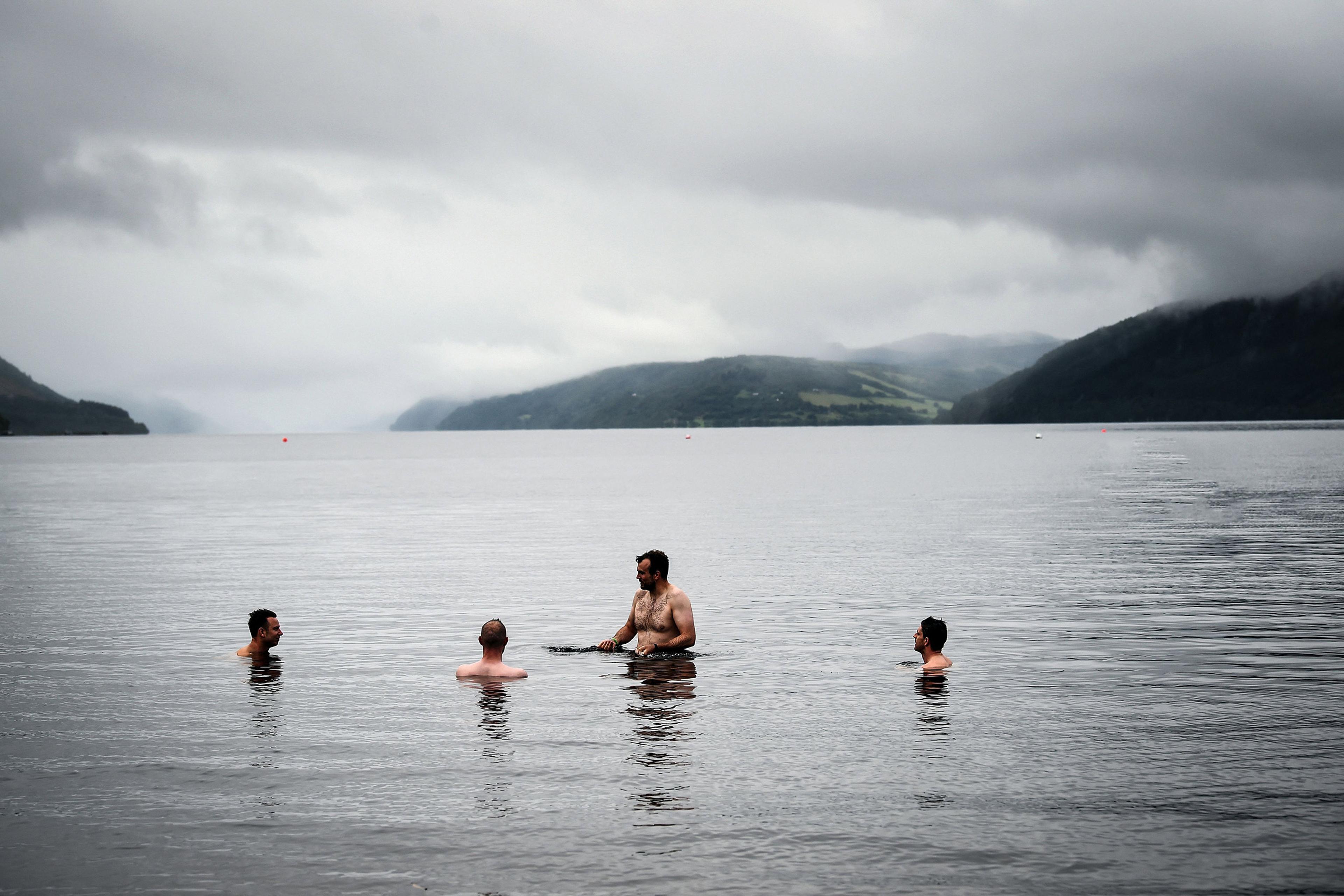 Four people swimming in a lake surrounded by foggy hills under a cloudy sky.