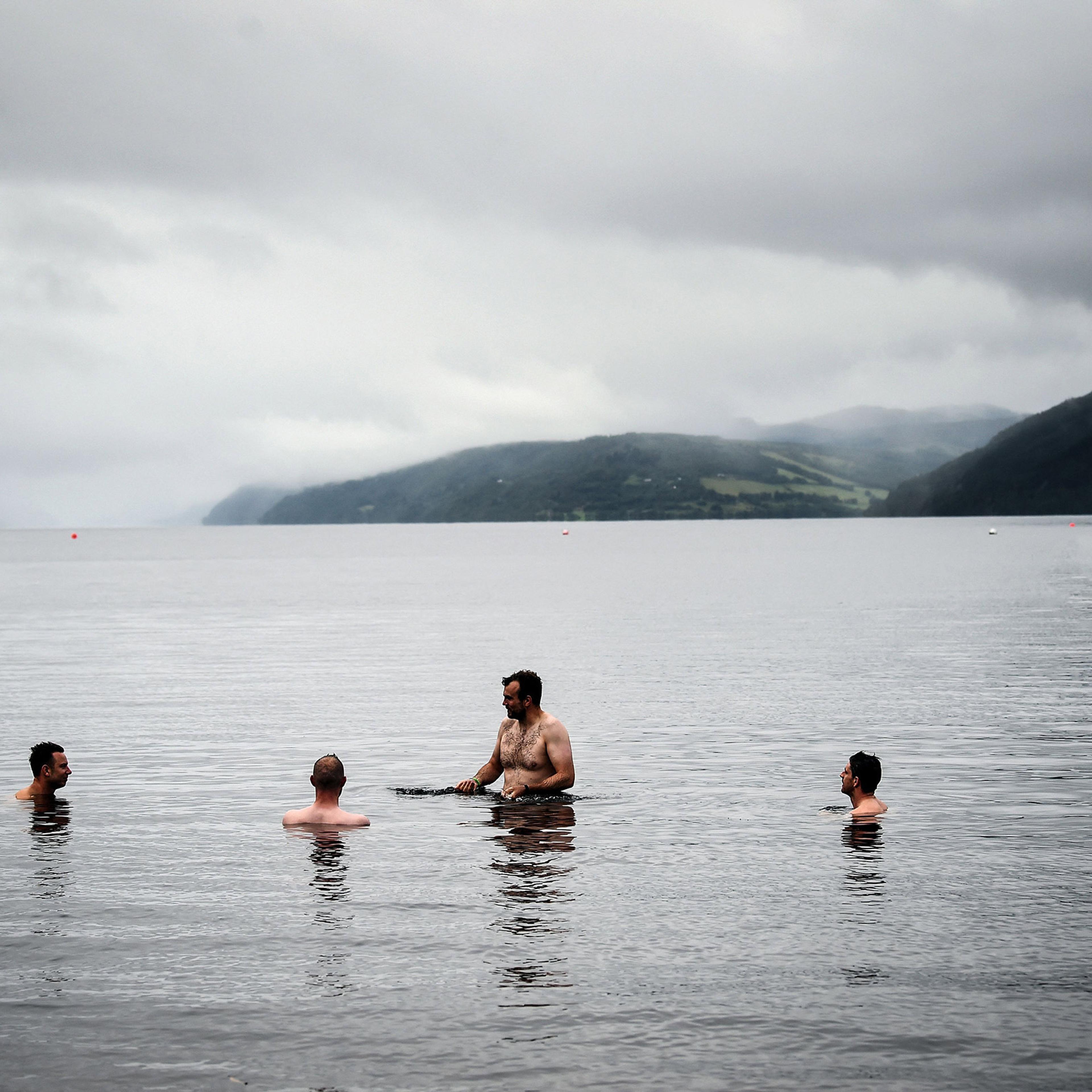 Four people swimming in a lake surrounded by foggy hills under a cloudy sky.