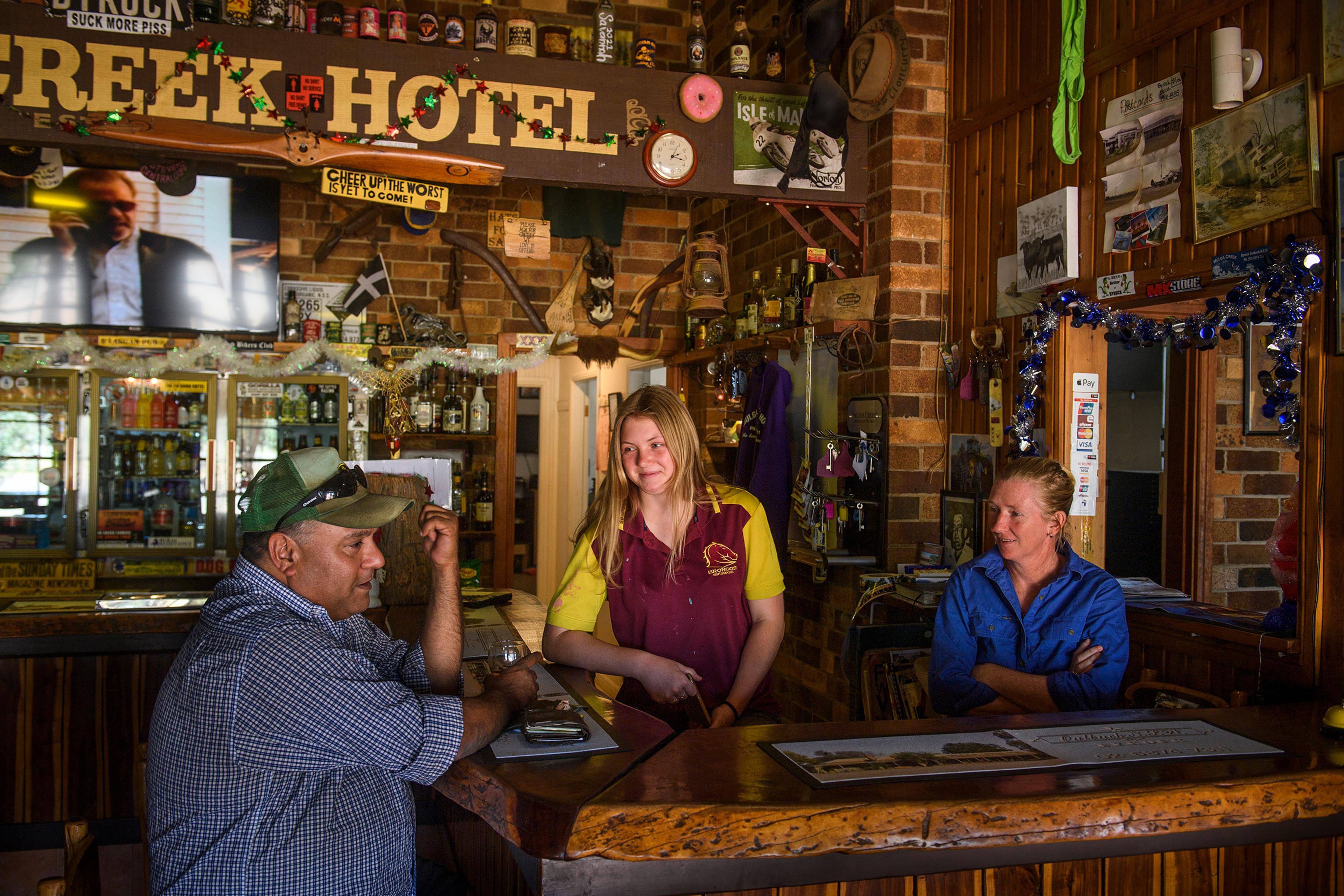 Three people chatting at a rustic bar in a pub, with a brick interior and various decorations.