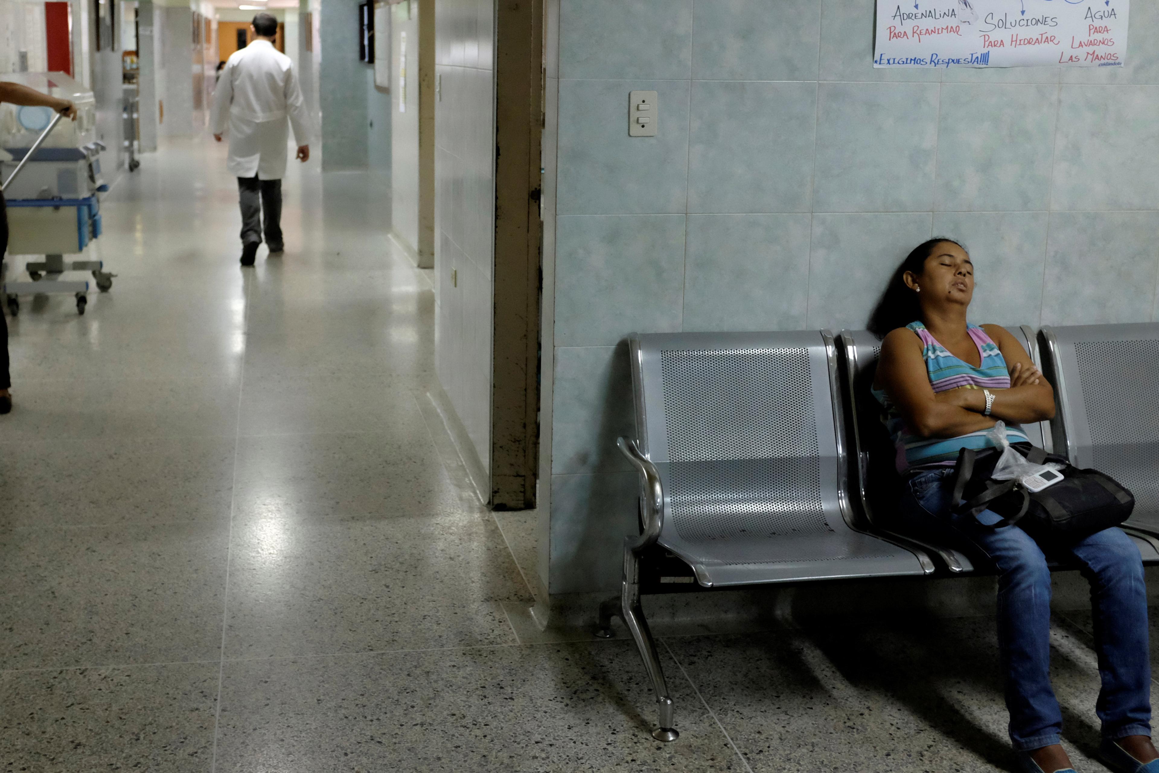 A hospital corridor with a woman asleep on a bench and a doctor walking away in the background.