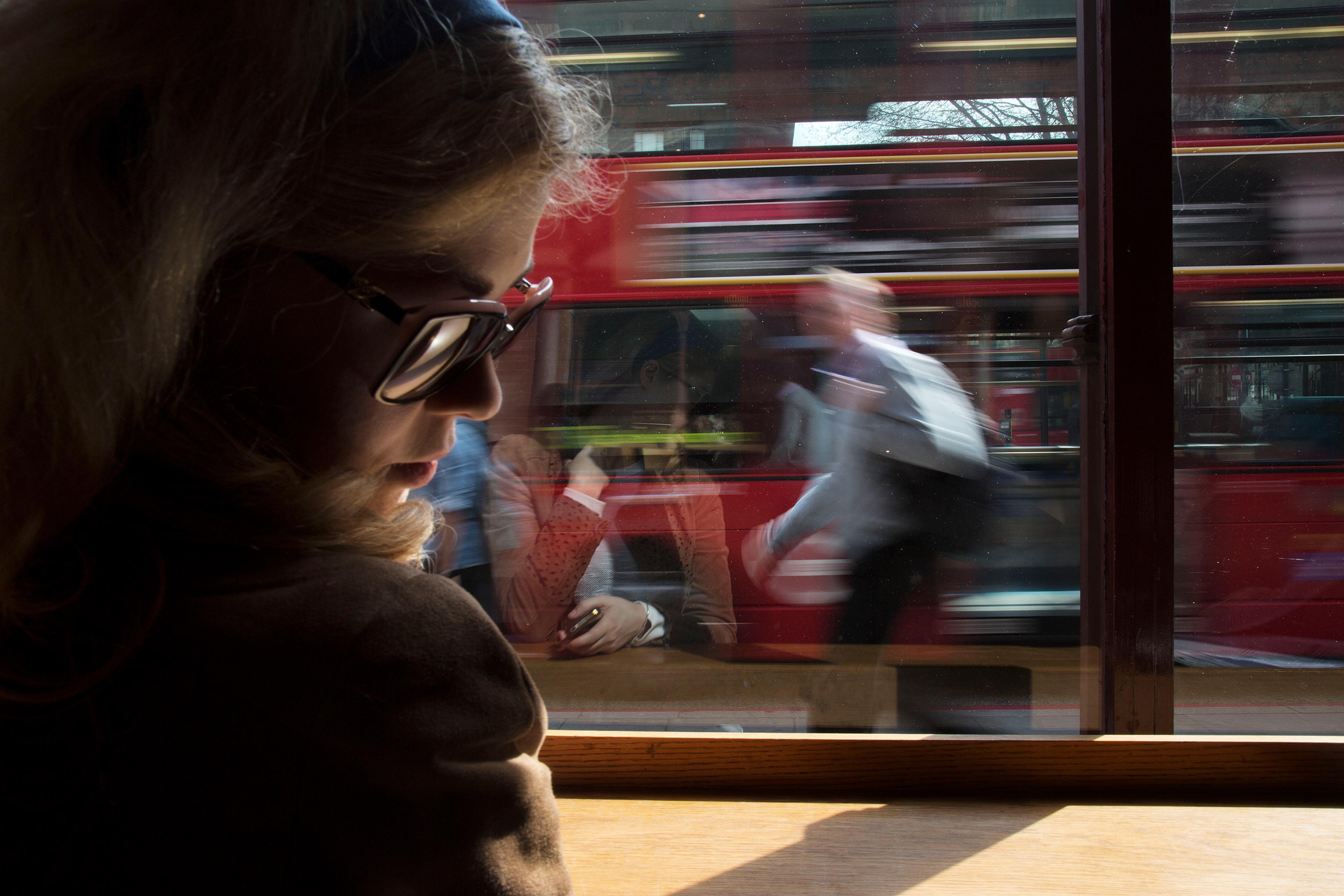 A woman inside wearing sunglasses, with a blurred reflection of people and a red bus moving outside the window.