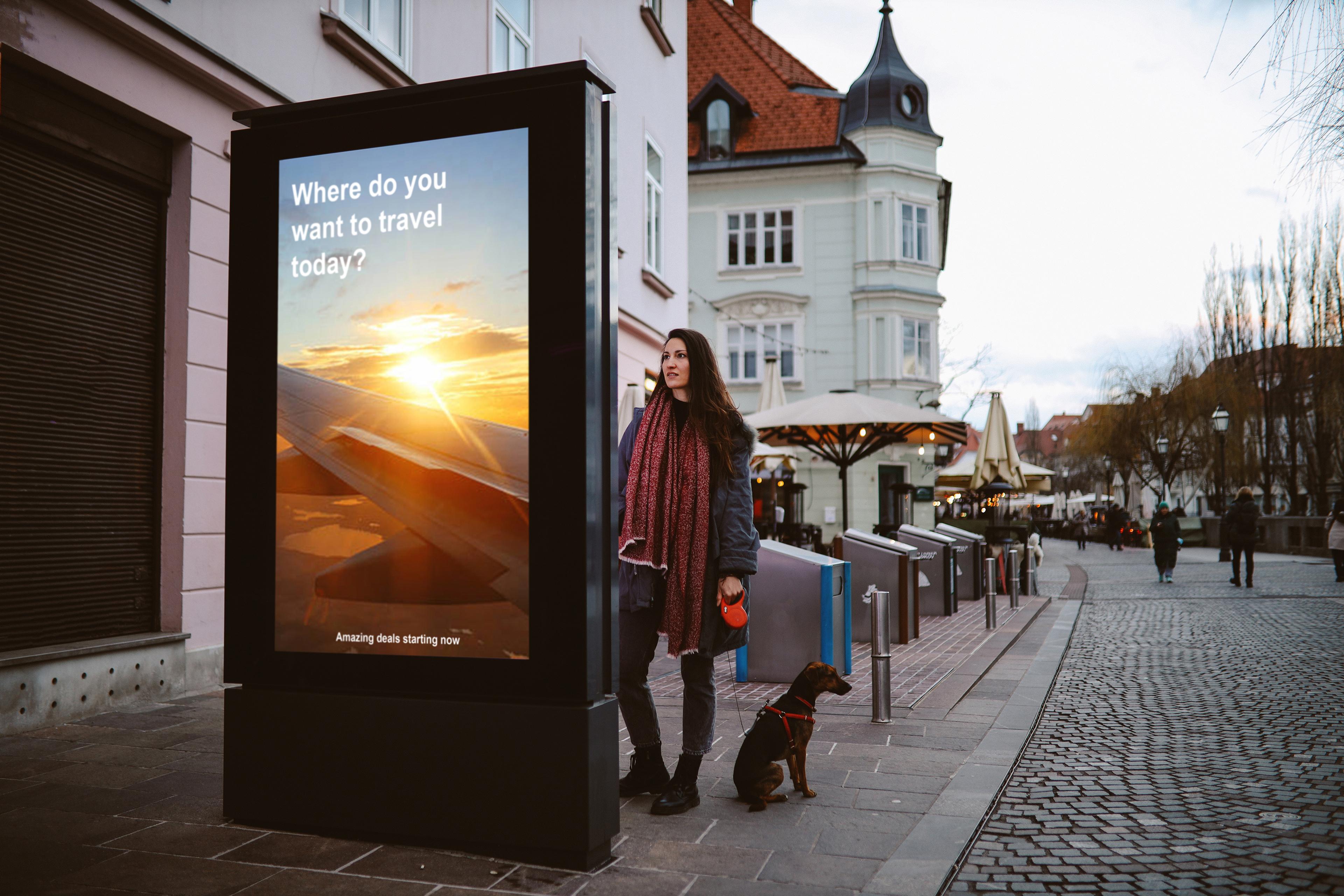 A woman and dog standing in a cobblestone street, by an outdoor travel advertisement showing a plane wing and sunset.