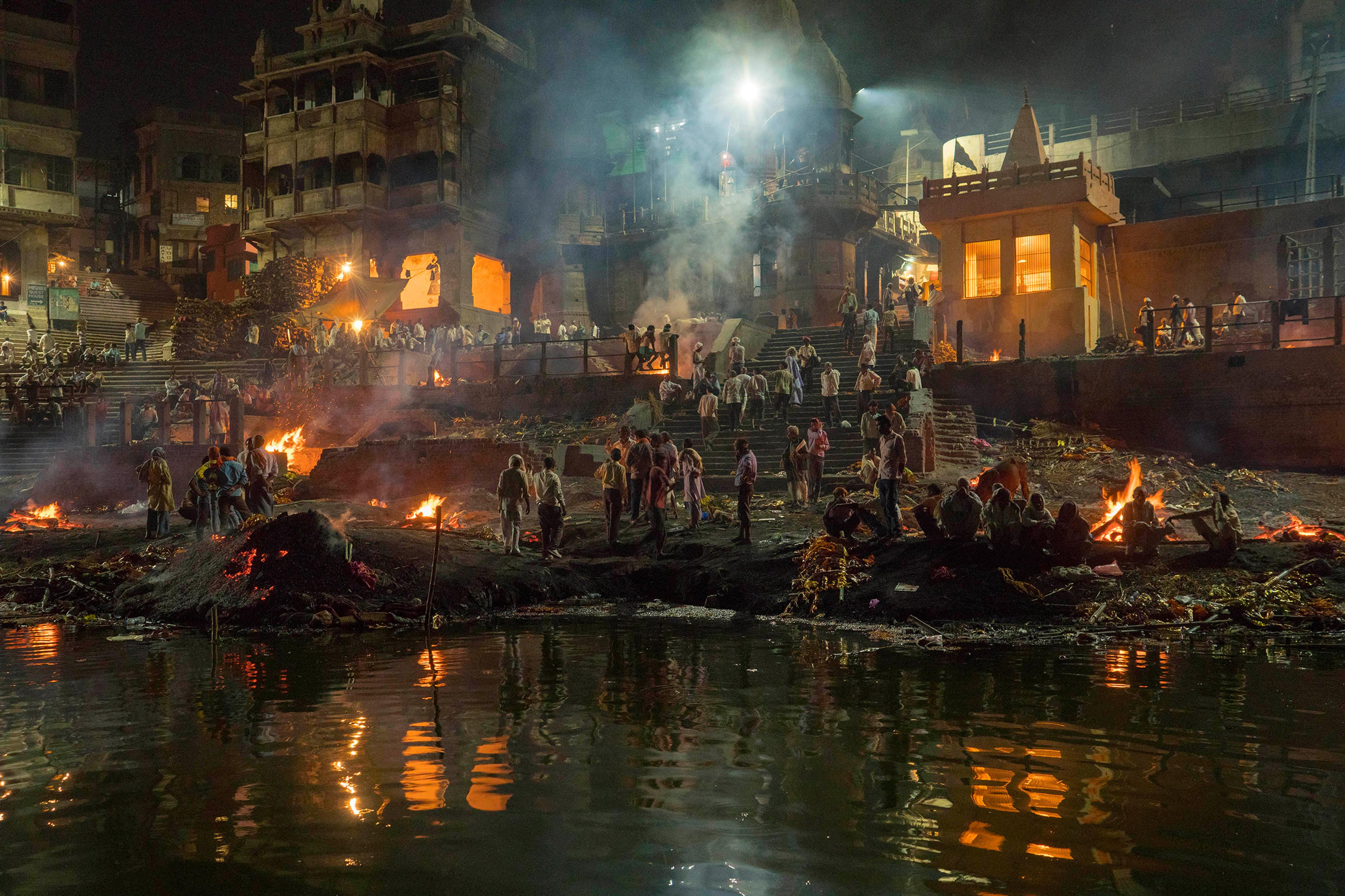 A cremation ceremony at night on a riverbank, surrounded by fires, people and historical buildings.
