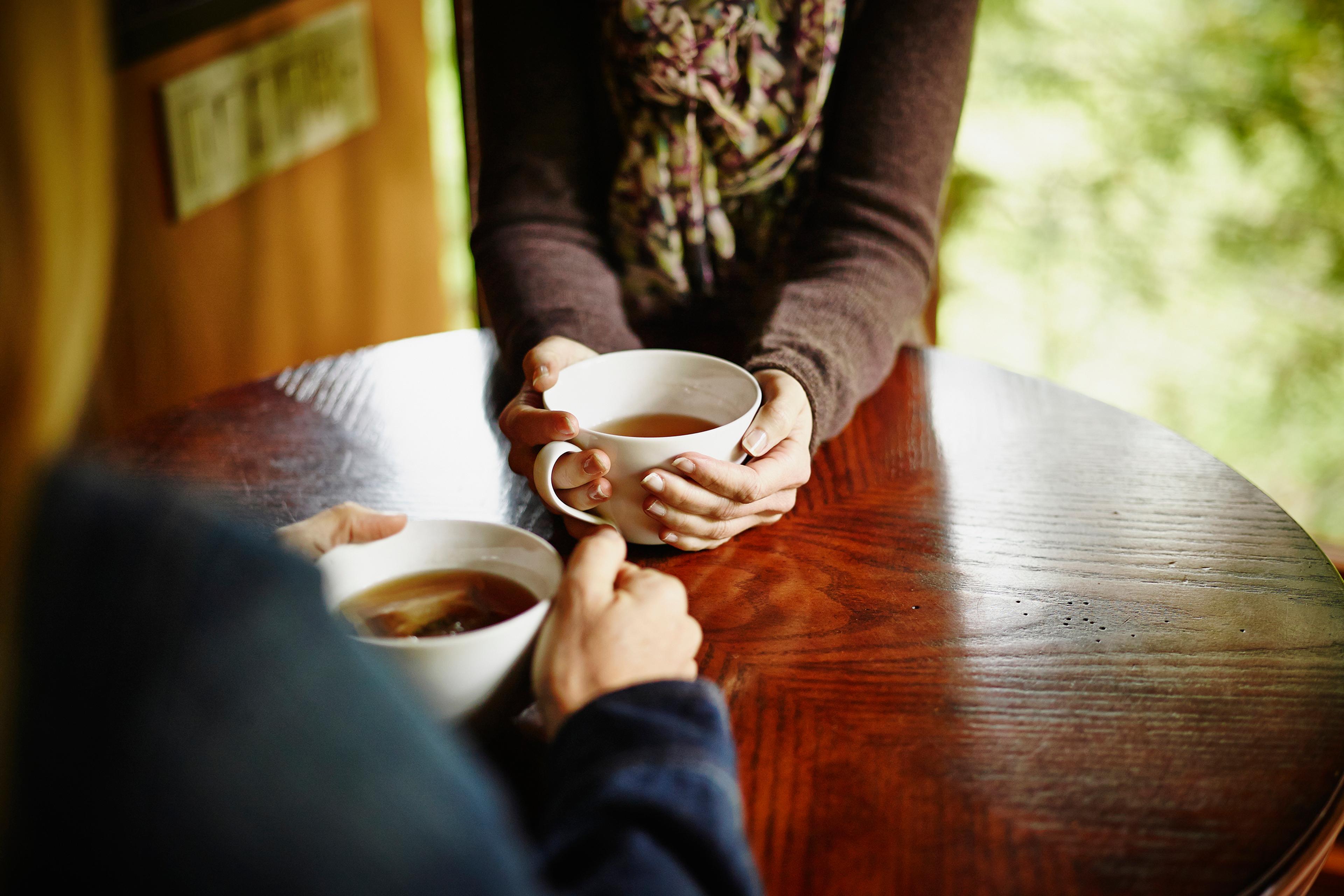 Two people holding mugs of tea at a round wooden table, one wearing a scarf, blurred green background.