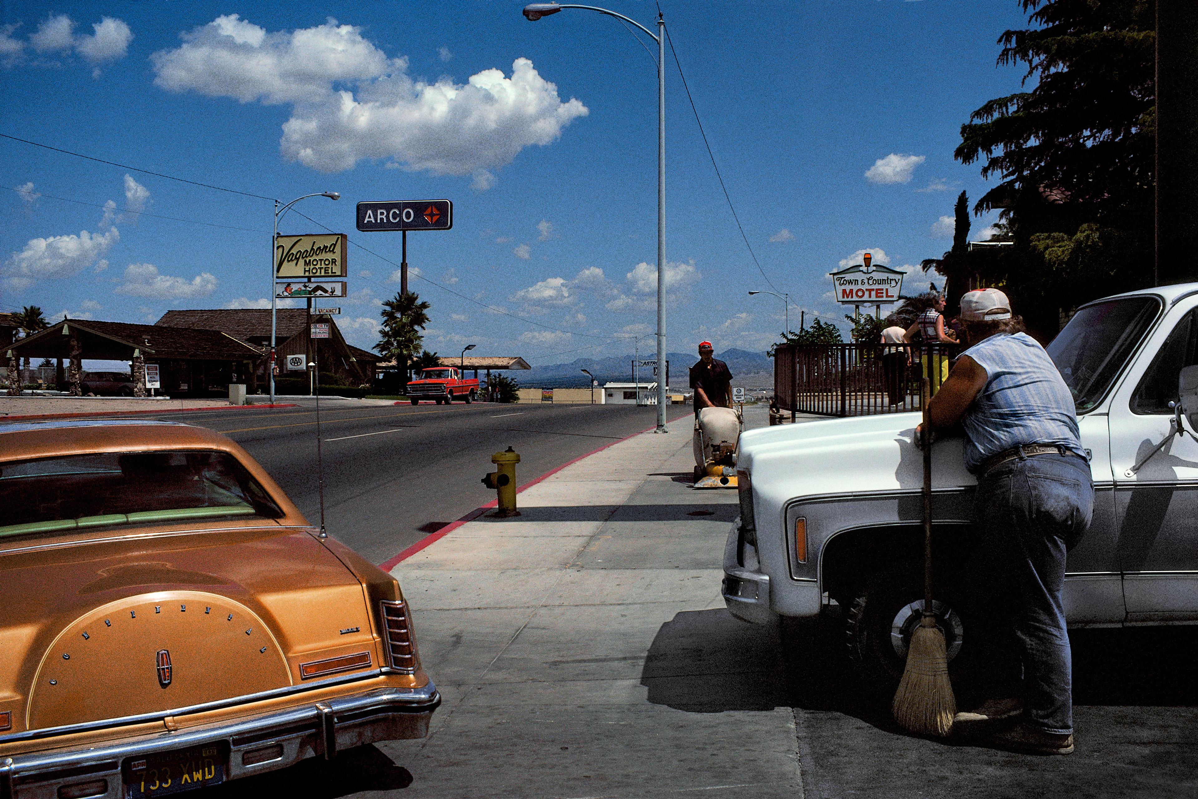 Photo of a street with vintage cars, motel signs and a person leaning on a truck holding a broom under a blue sky.