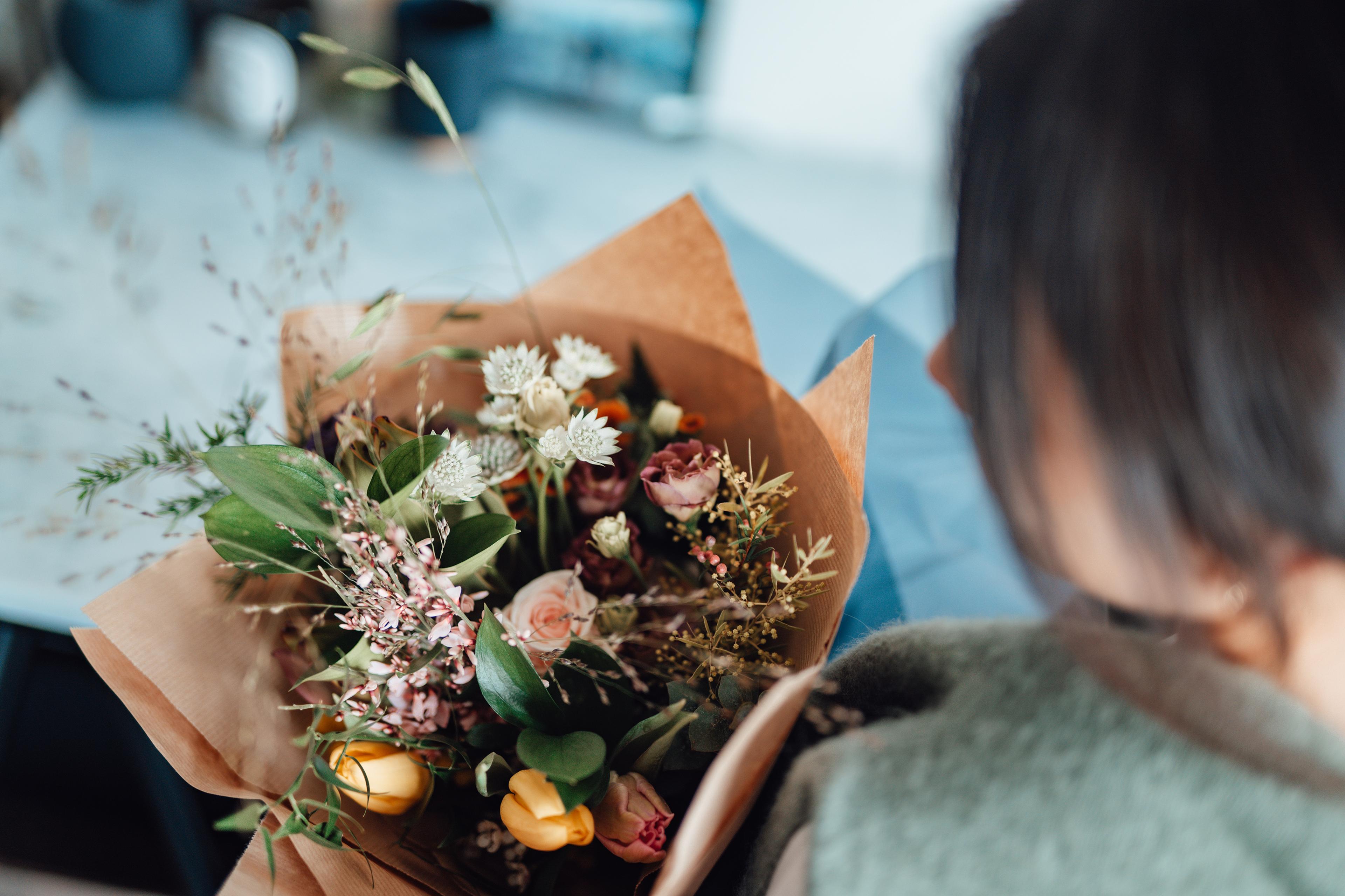 A photo showing a person holding a bouquet of mixed flowers wrapped in brown paper, viewed from above and behind.